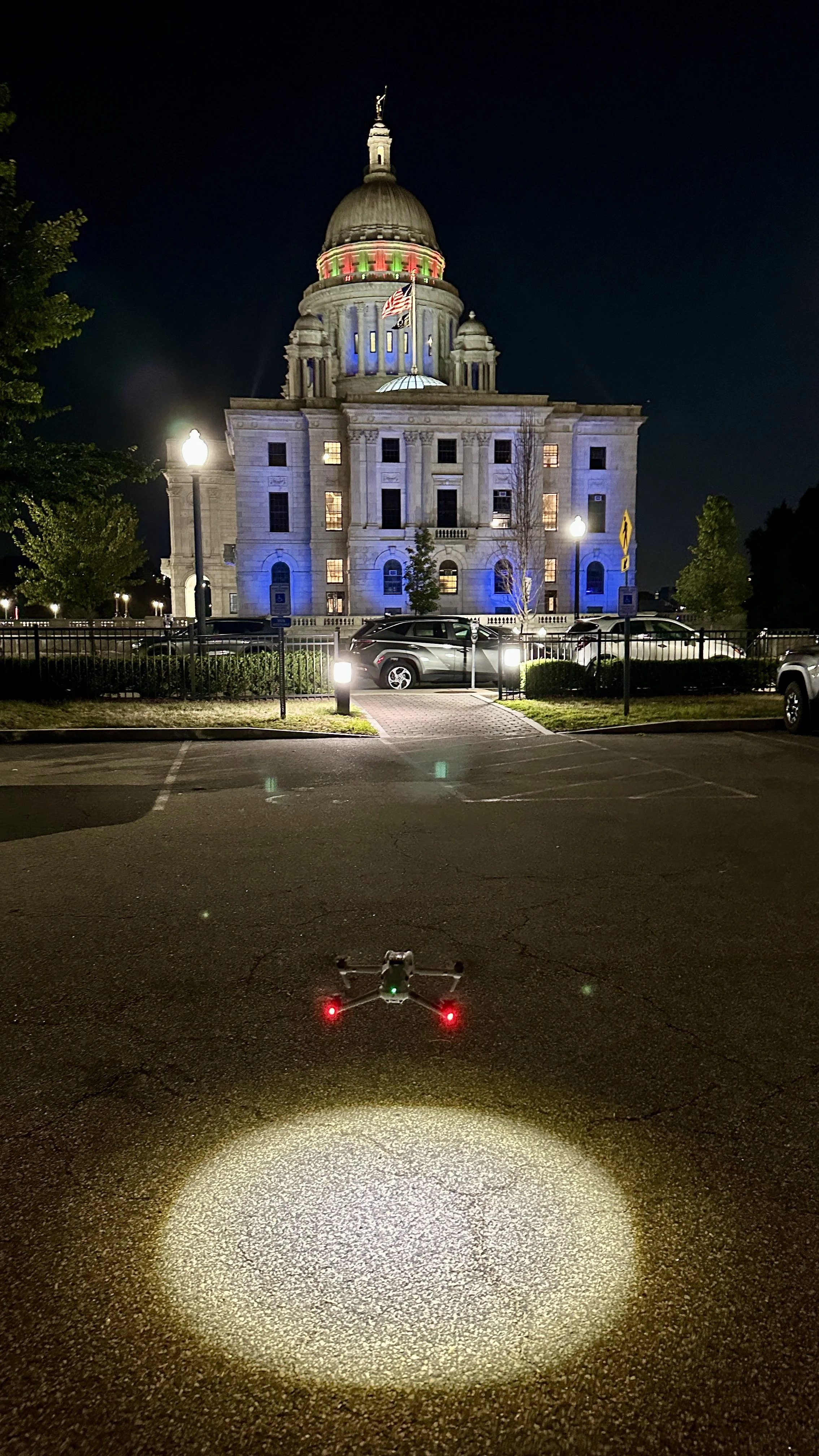A drone with the Rhode Island State House in the background illuminated at night.