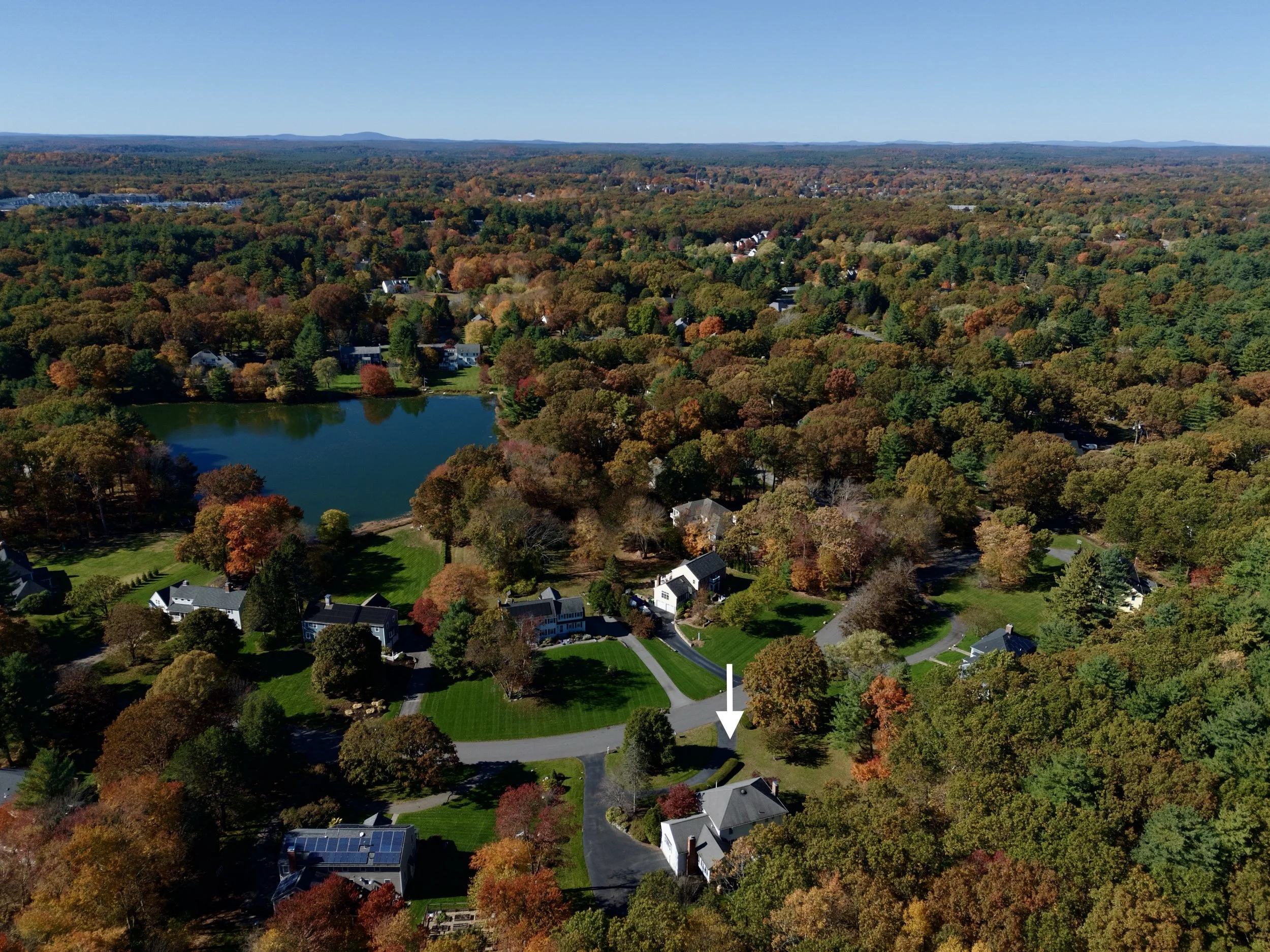 Real estate aerial, drone photography of a house in Sudbury, Massachusetts in a residential neighborhood with blue skies, a lake and foliage near Boston.