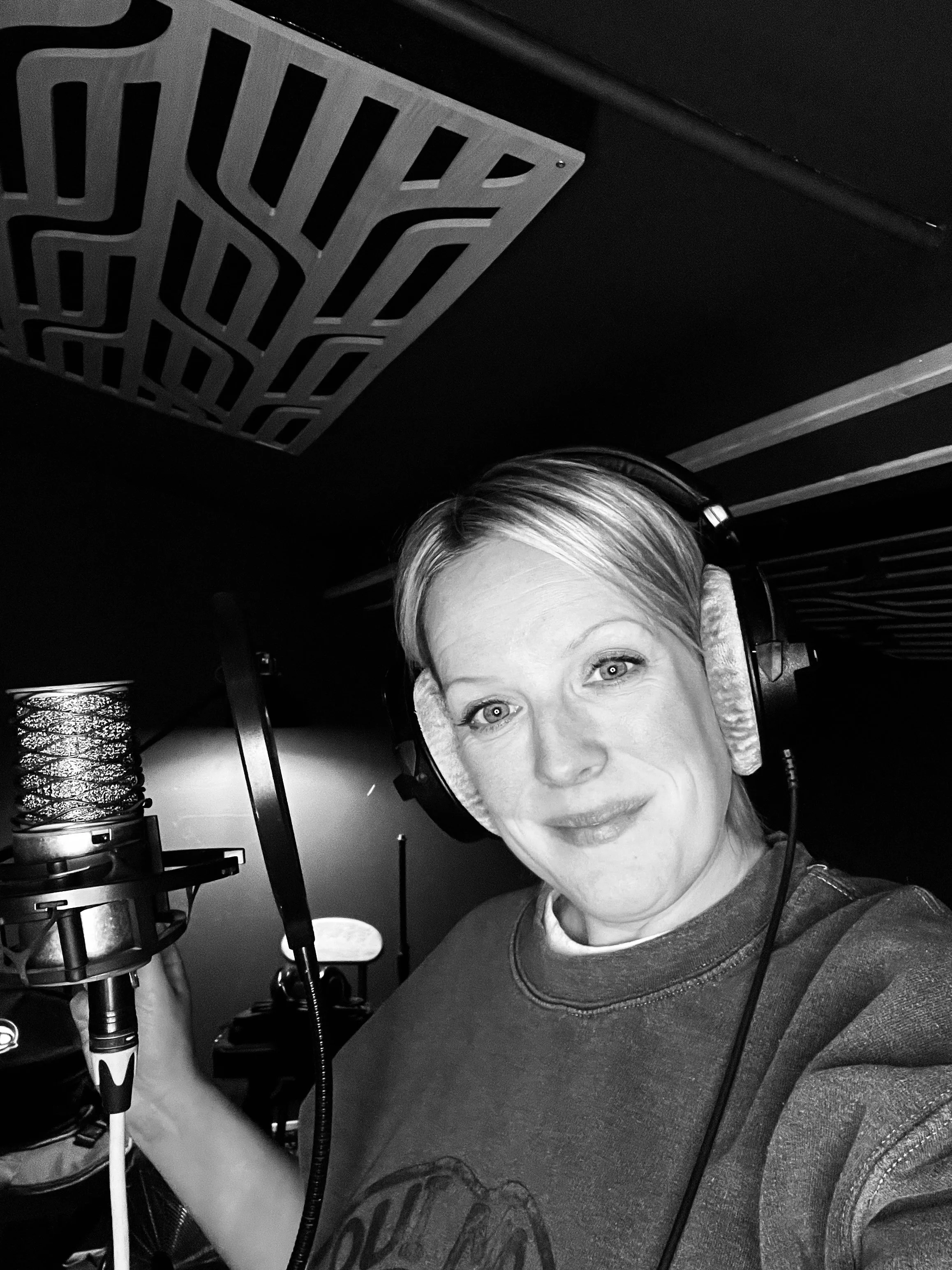 A woman in a recording studio wearing headphones and holding a microphone, smiling at the camera in black and white.