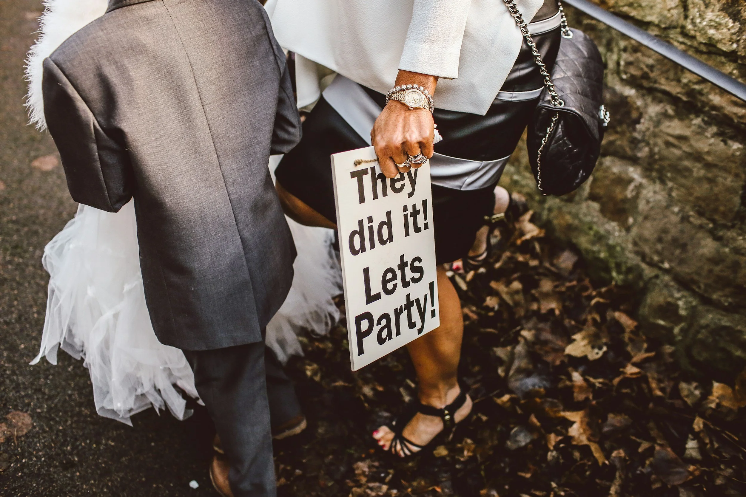 A close-up of two people standing outdoors. One person is holding a sign that reads 'They did it! Let's Party!' while wearing a watch, rings, and a bracelet. The person is dressed in black and white, with a black handbag hanging from their shoulder. The other person is dressed in a gray suit with a tutu visible underneath, and brown shoes. The surroundings include fallen leaves and a stone wall.