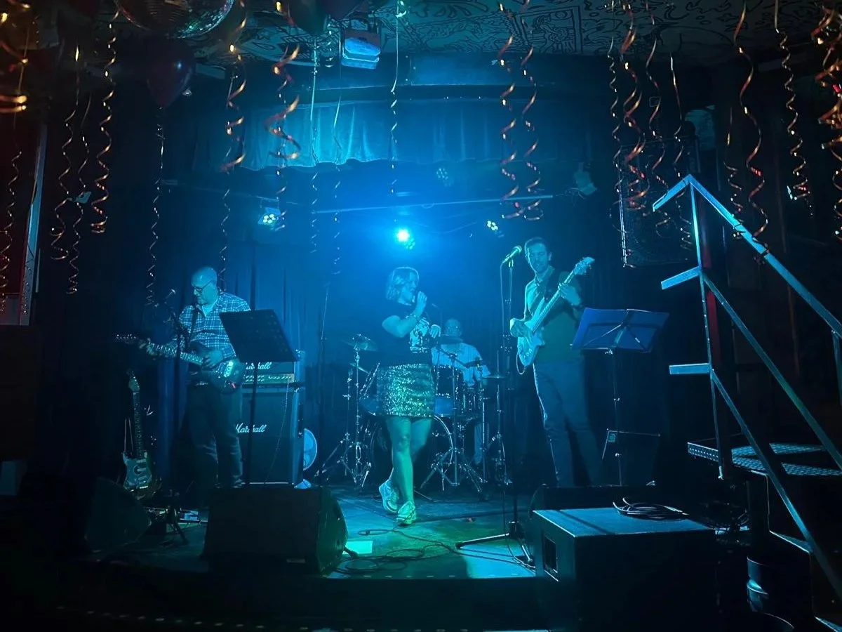 A band performing live on stage with four musicians, including a female singer, under blue stage lighting, surrounded by musical instruments and equipment, in a dimly lit venue with hanging decorations.