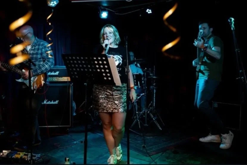 A band performing on stage with a female singer and two male musicians, one playing guitar and the other playing saxophone, in a dimly lit venue.