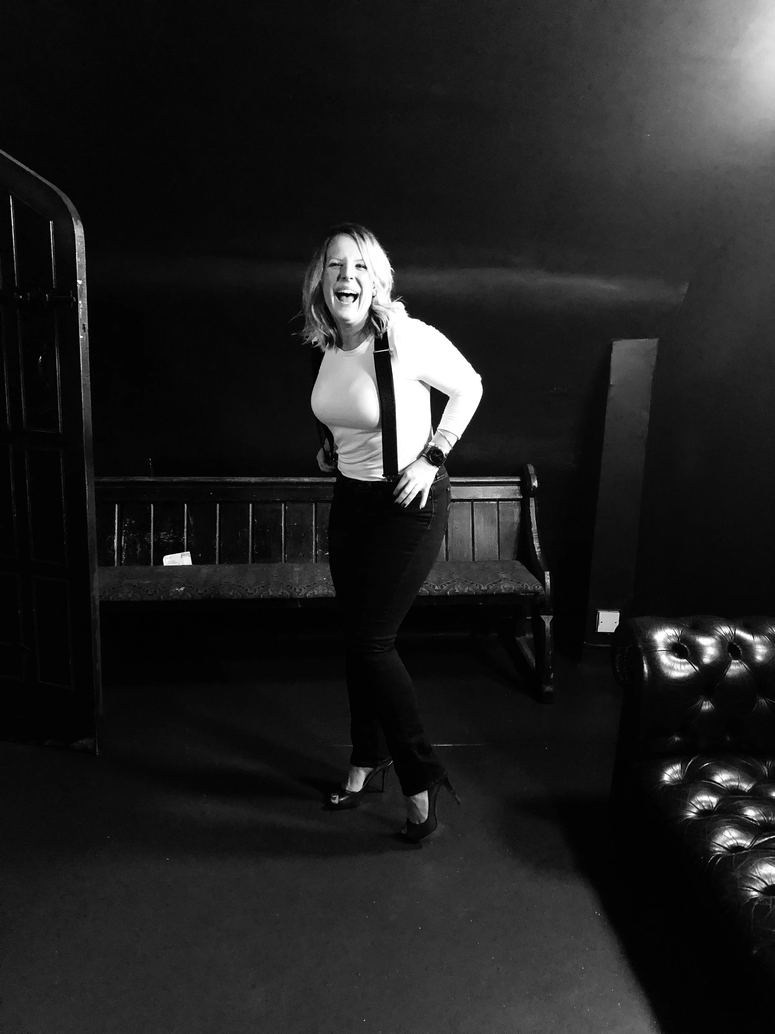 A woman laughing and posing in black and white, wearing a white top, black pants, high heels, and suspenders. She is standing in a room with dark walls, a wooden bench, and a leather armchair.