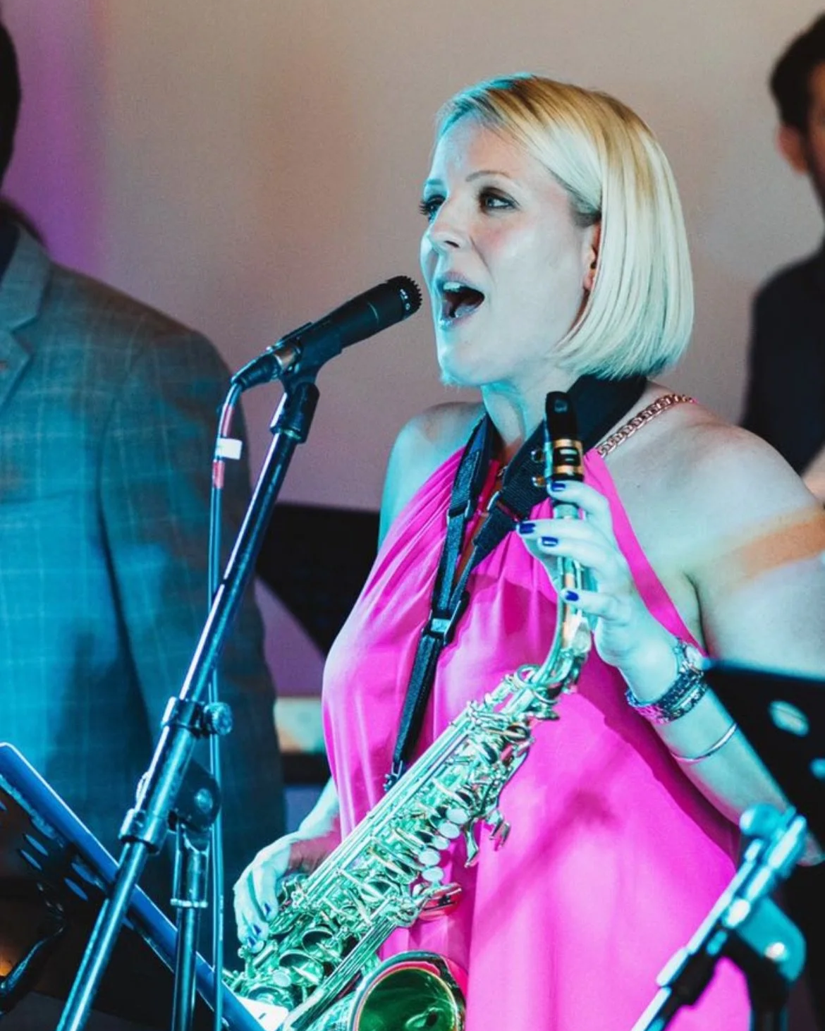 A woman singing into a microphone while holding a saxophone, dressed in a pink dress.