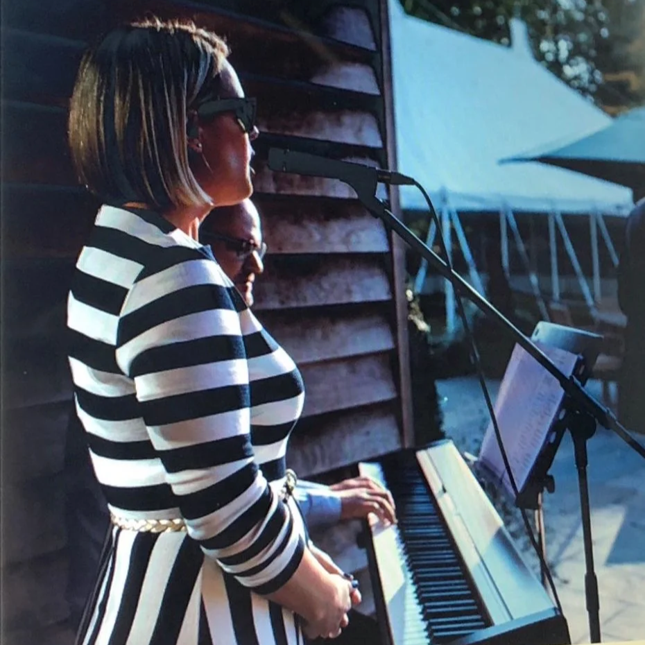 A woman singing next to a man playing a piano outdoors. The woman is in a navy and white striped dress and sunglasses, with a music stand in front of her, surrounded by wedding marquees.