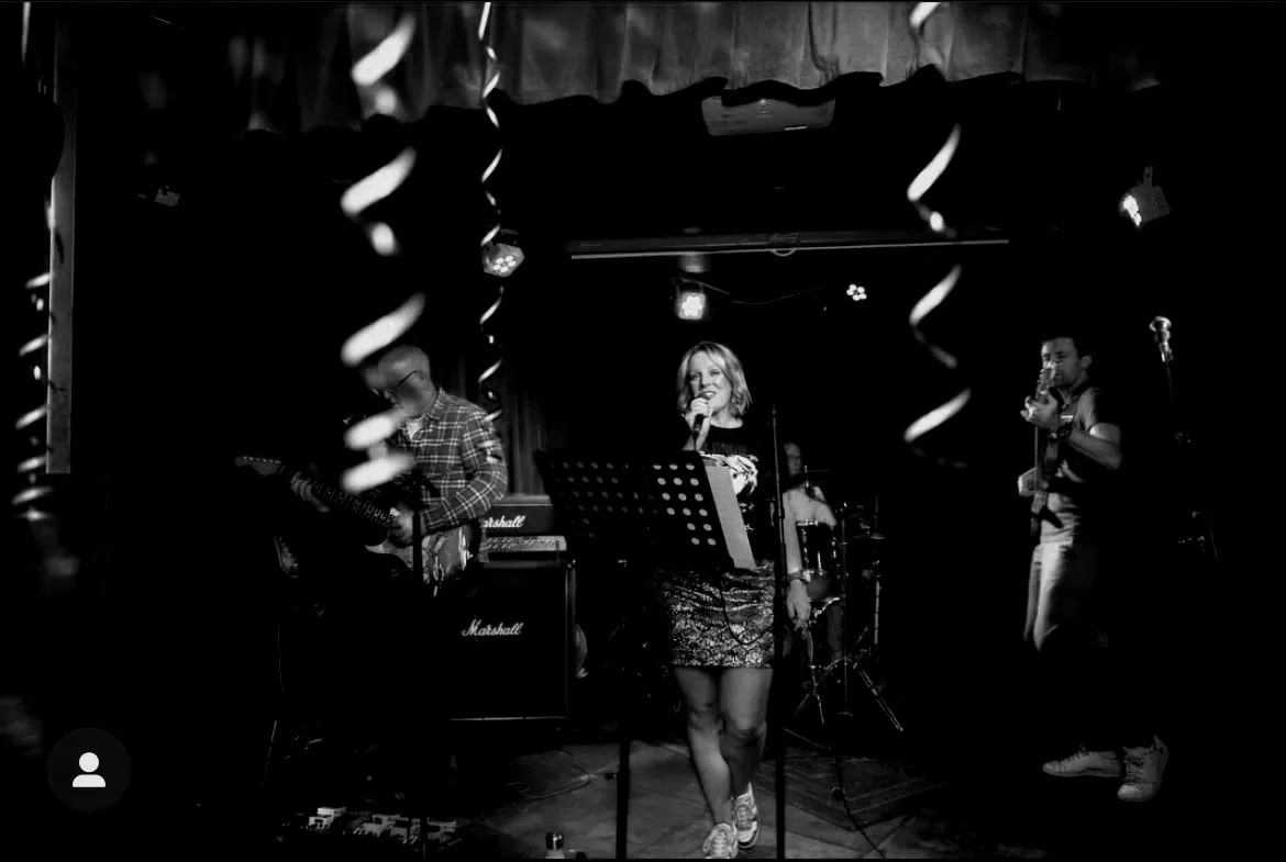 Black and white photo of a live music performance with a band on stage. A woman is singing into a microphone, standing at the center. To her left is a guitarist, and to her right is a bass guitarist
