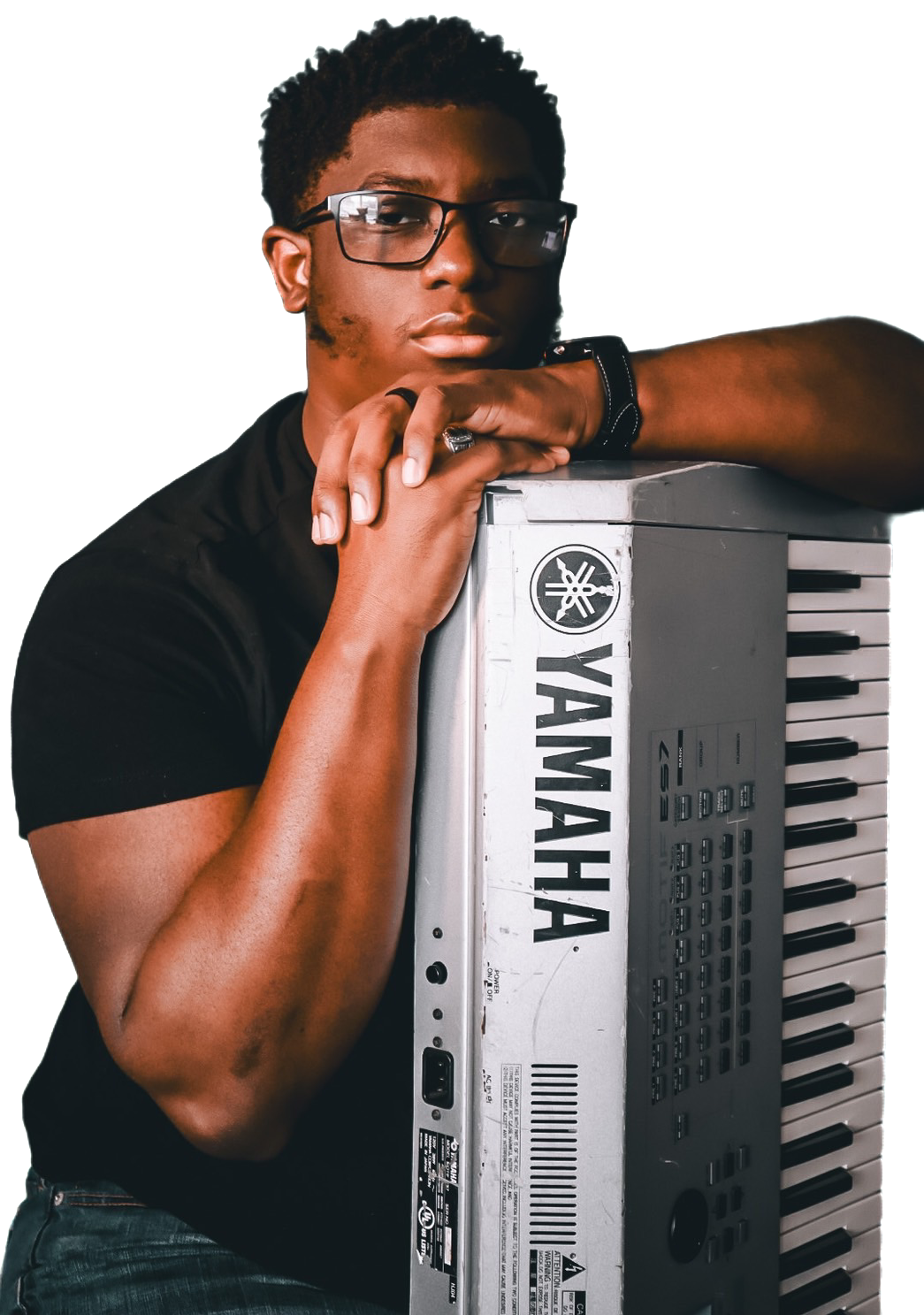 A young man wearing glasses and a black shirt, leaning on a Yamaha keyboard, with a serious expression.