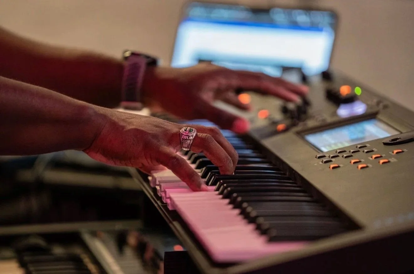 Close-up of hands playing a keyboard synthesizer, with a computer screen in the background.