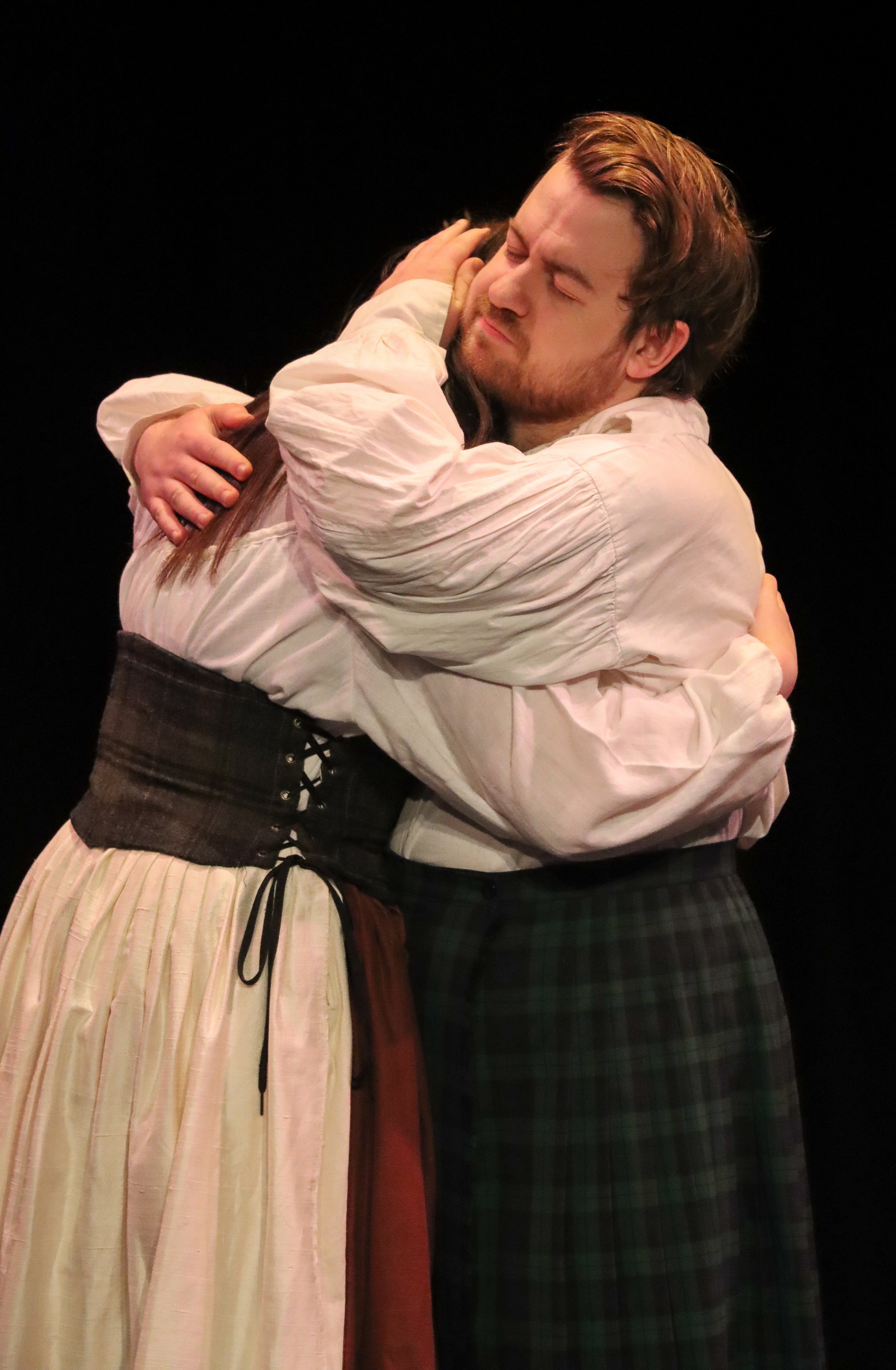 Two actors embracing on stage, dressed in period costumes, with a black background.