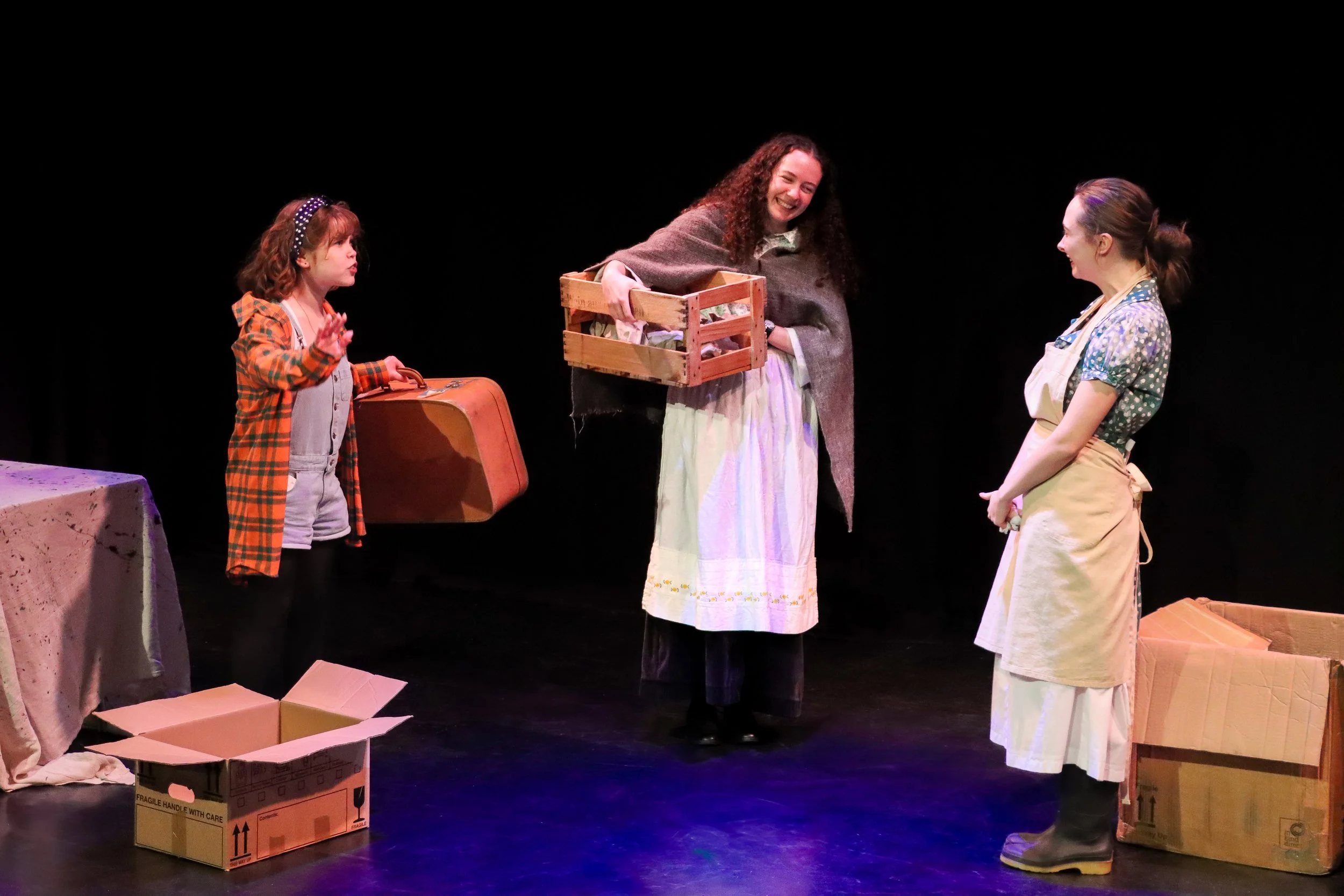 Three women on stage, one young girl and two adult women, engaged in a theatrical performance involving cardboard boxes and a suitcase, with a black background.