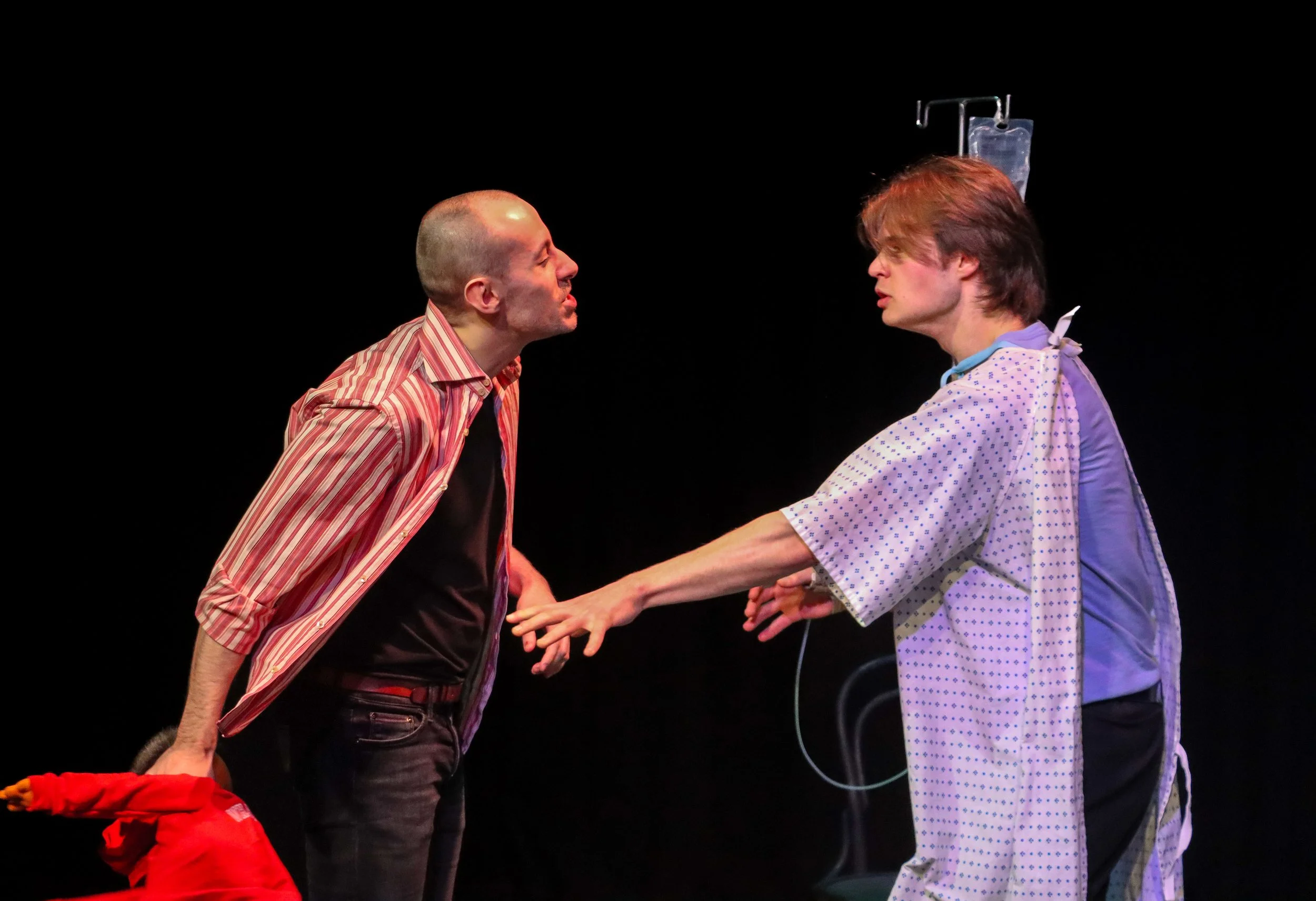 Two men are engaged in a theatrical confrontation on a dark stage. One man, bald and wearing a striped shirt, leans towards the other, who has shoulder-length hair and wears a hospital gown, with an IV stand behind him. The man in the gown extends his arm toward the other.