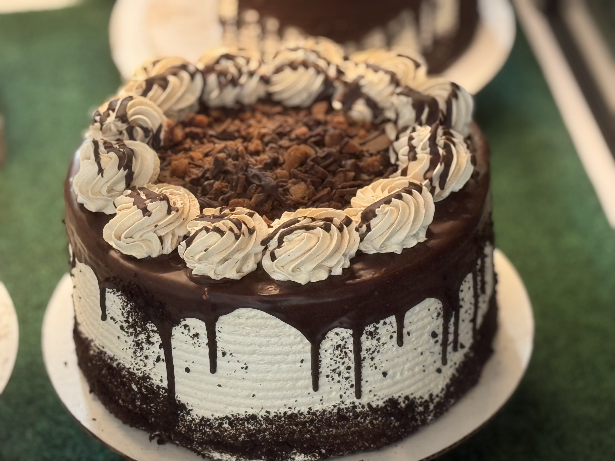 Chocolate cake with cream frosting, chocolate drizzle, and whipped cream rosettes, topped with chocolate shavings, on a green surface.