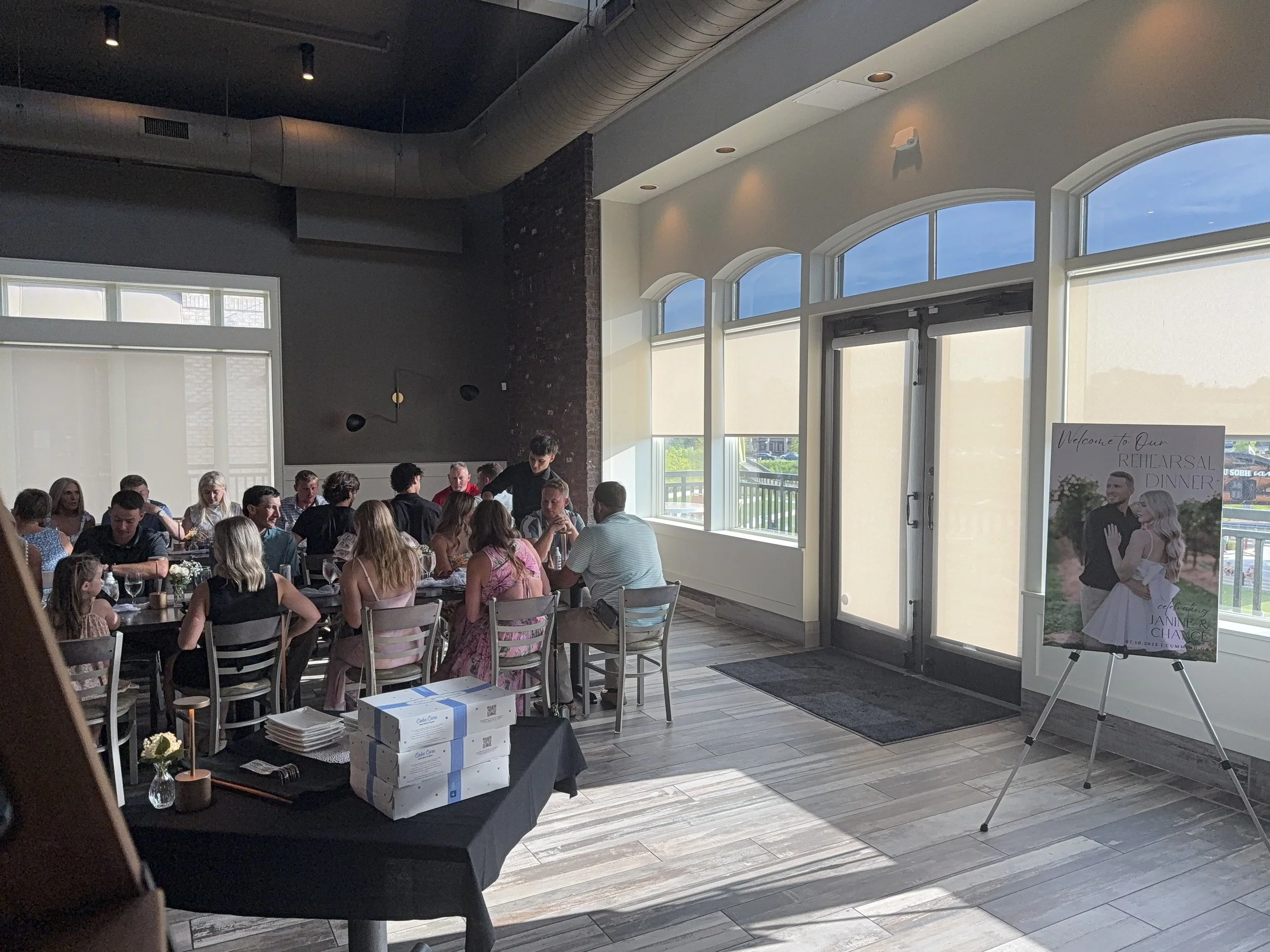 A gathering of people seated at tables inside a bright, modern event space with large windows, a welcoming poster, and a table with boxes and plates near the entrance.