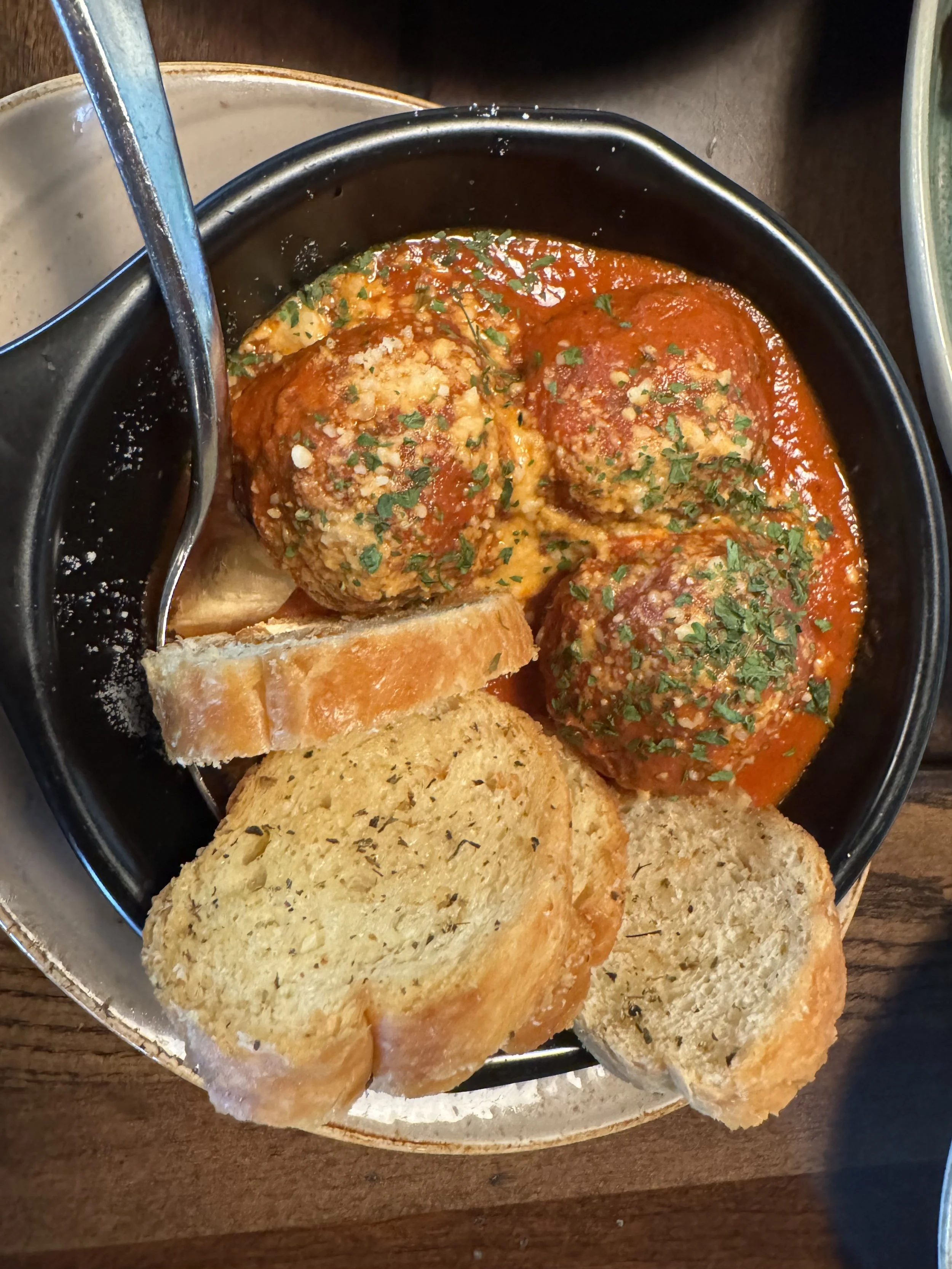 A bowl of Italian meatballs in tomato sauce with chopped herbs, served with slices of toasted garlic bread and a slice of bread on the side.