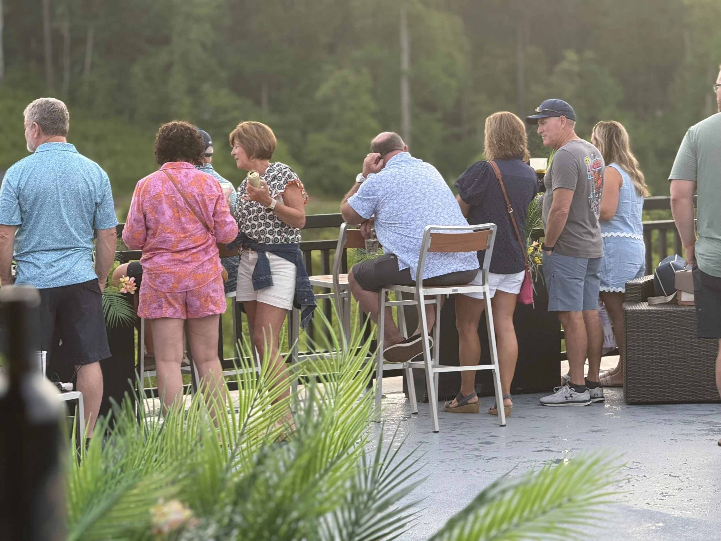 A group of people gather on an outdoor patio, socializing and enjoying drinks, with a lush green landscape in the background.
