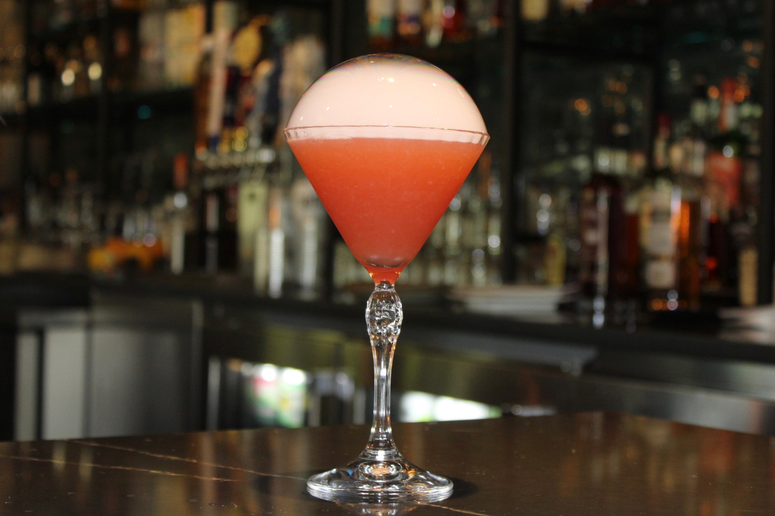 A cocktail glass with a layered pinkish-red and white beverage, topped with a white foam, sitting on a dark bar counter with blurred shelves of bottles in the background.