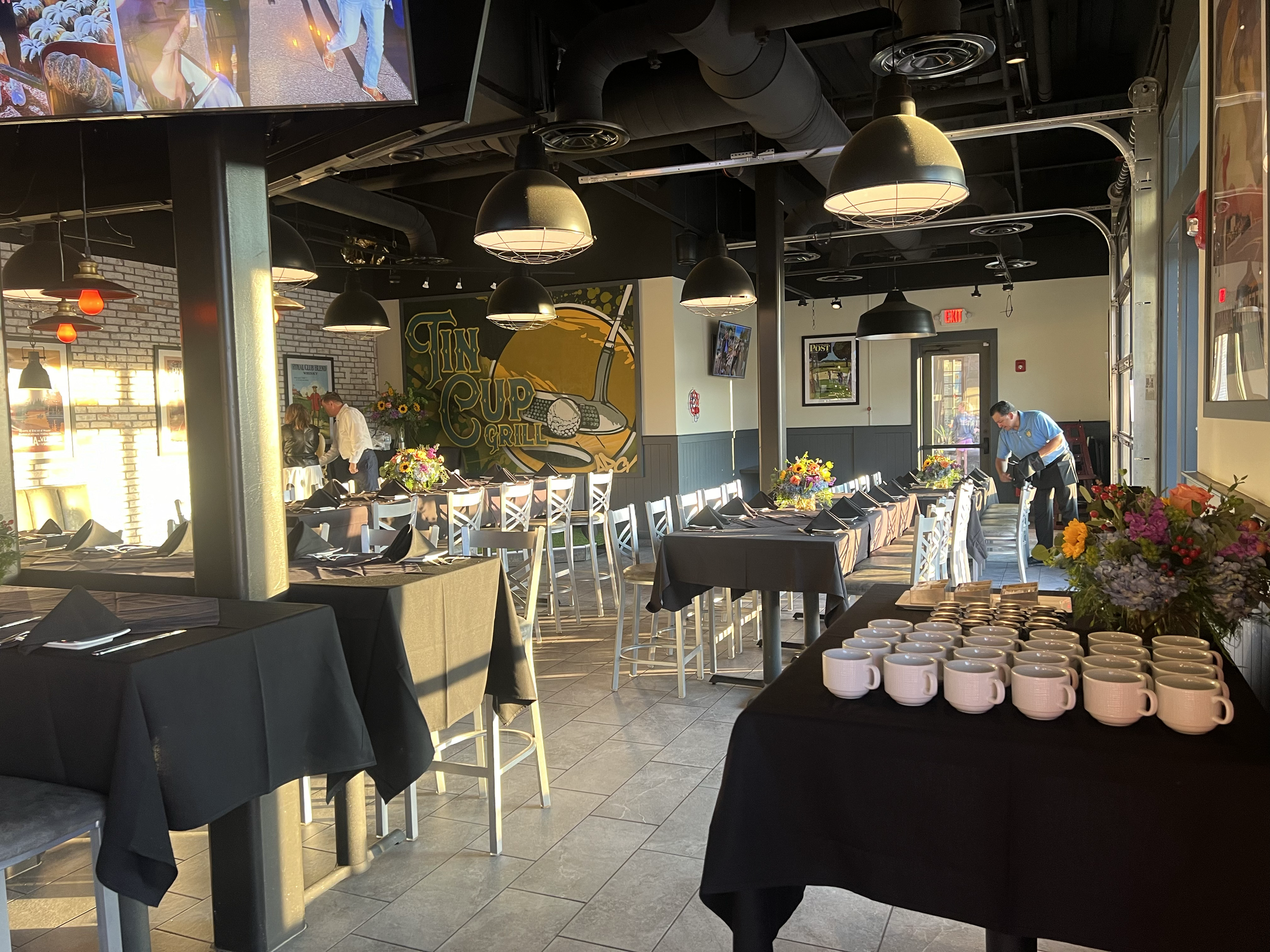 Interior of a restaurant set up for an event, with tables covered in black tablecloths, black napkins, and floral centerpieces. Coffee mugs are arranged on a table near the window. Staff members are preparing the space, and sunlight streams through t