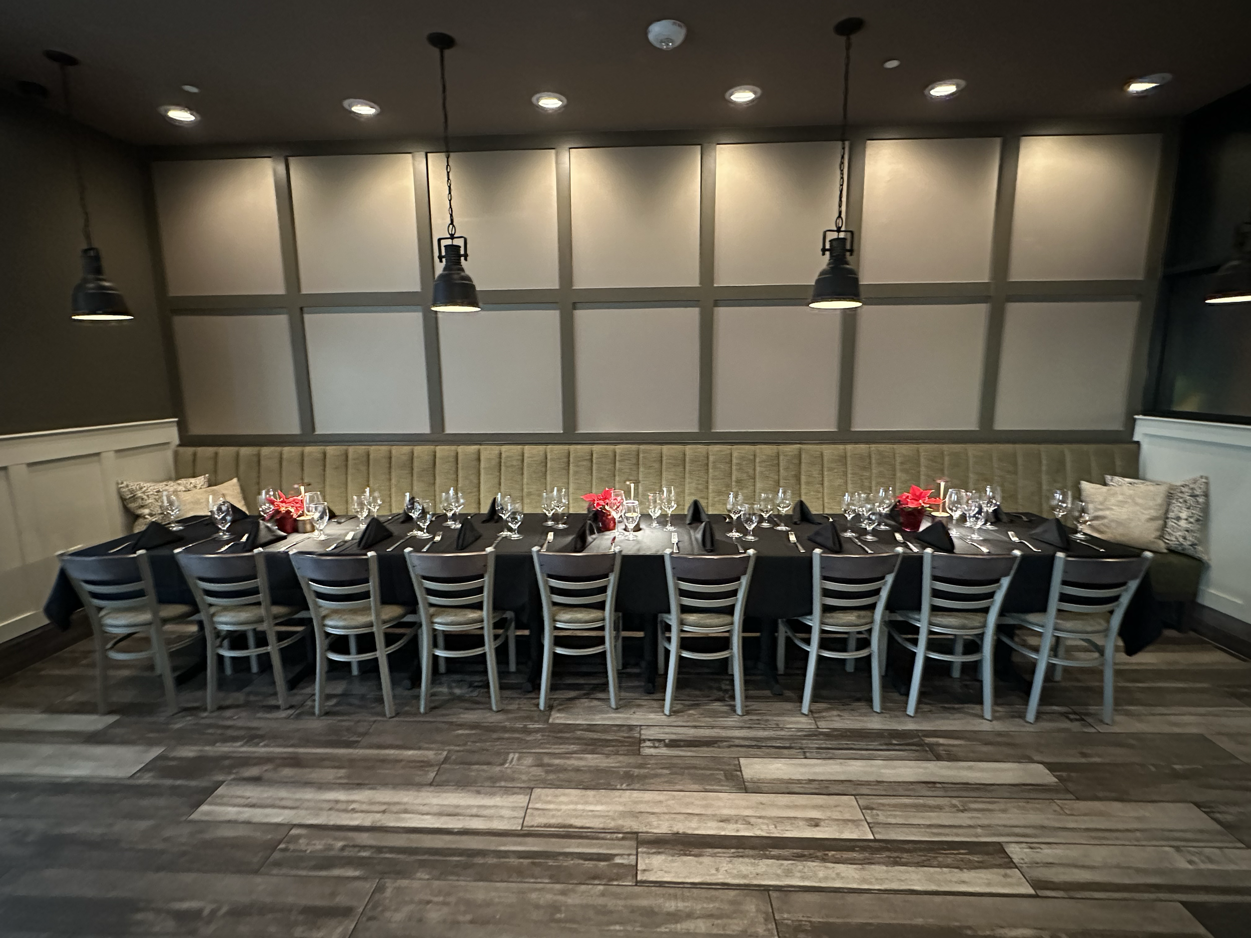 Long rectangular dining table set for a formal meal with black tablecloths, folded black napkins, wine glasses, water glasses, and small floral centerpieces in red pots, surrounded by chairs and a cushioned banquette with pillows against the wall, in