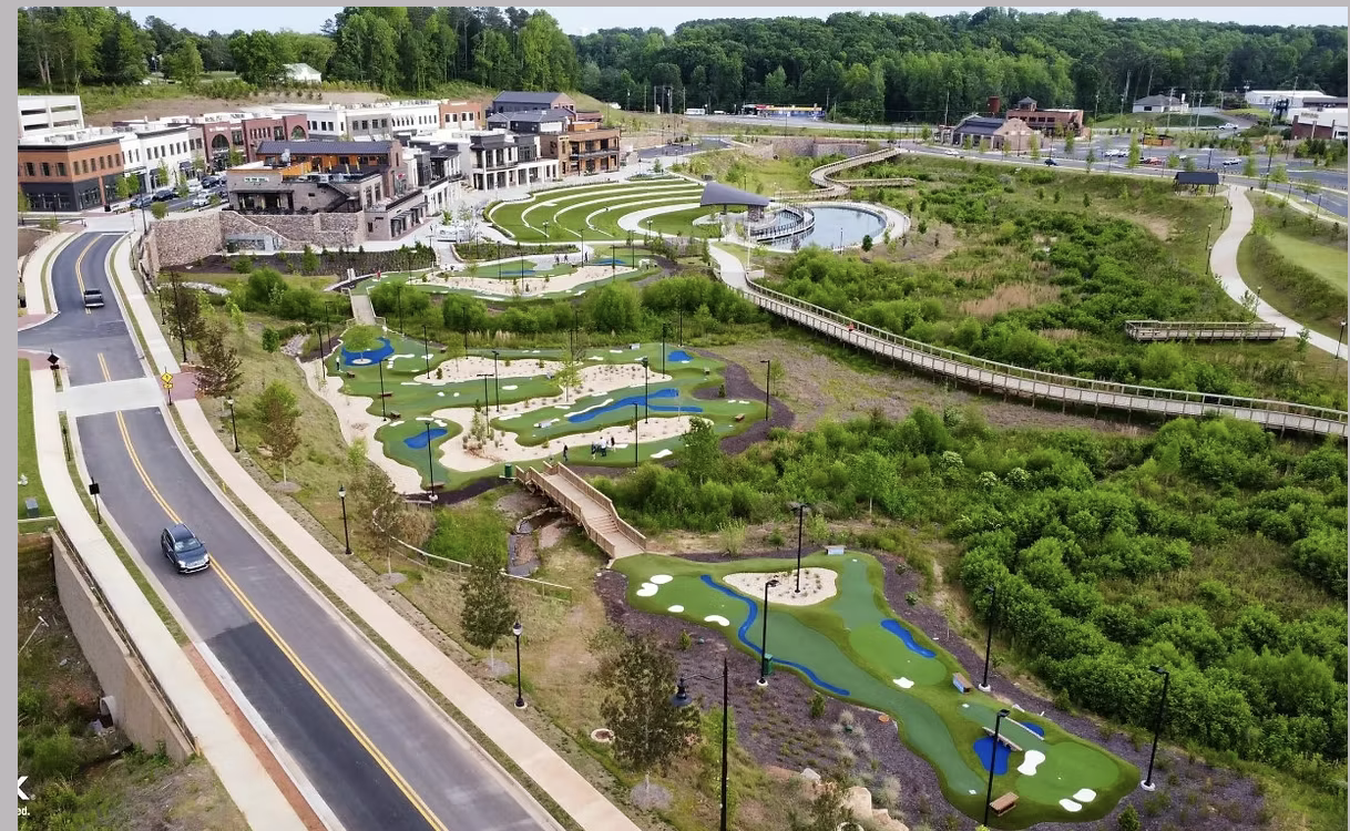 Aerial view of a mini golf course with multiple holes, water features, and scenic landscaping, with a series of commercial buildings and roadways in the background.