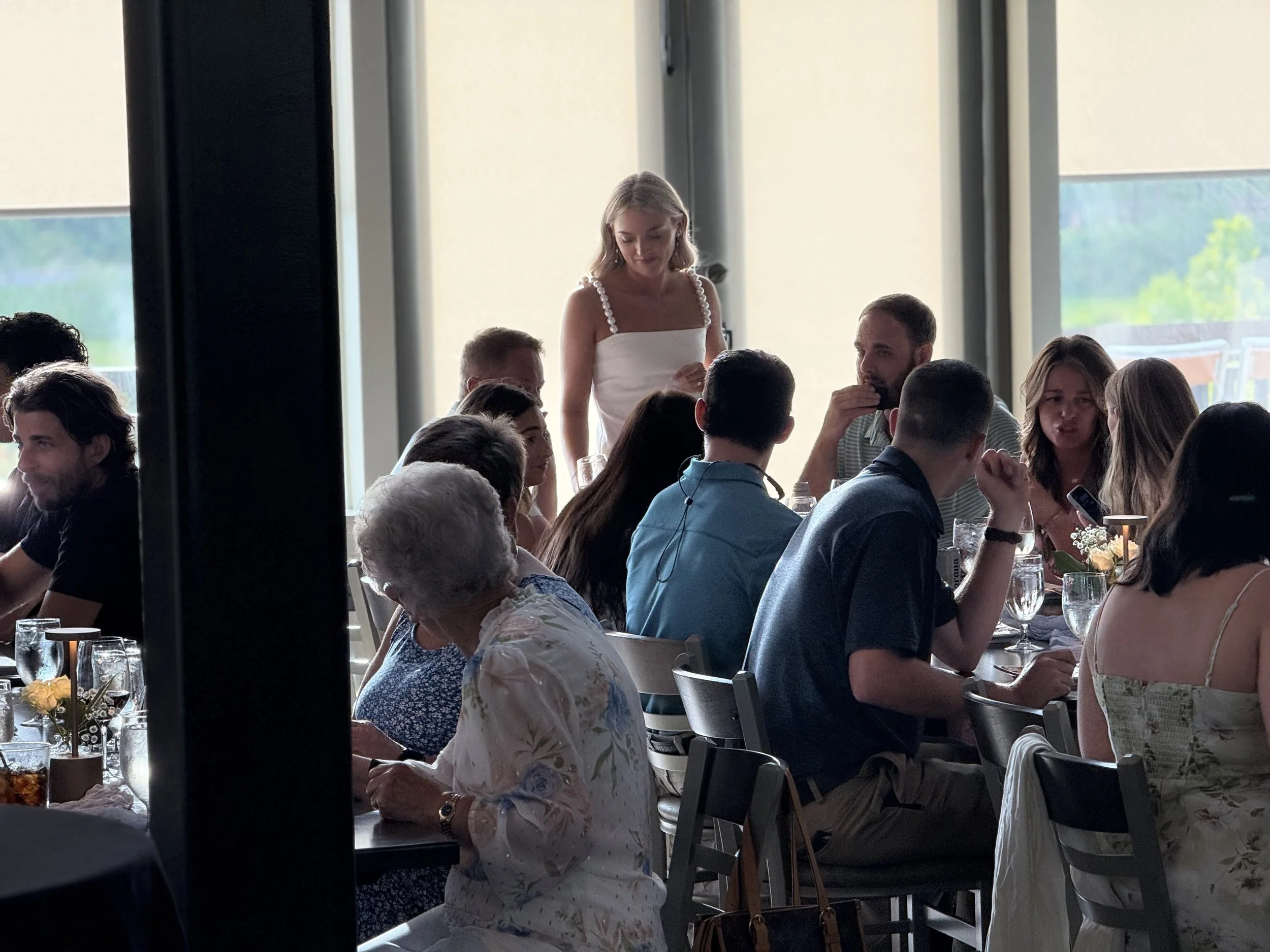 People gathered around a dining table during a celebration, with a woman standing and speaking to the group in a well-lit room.