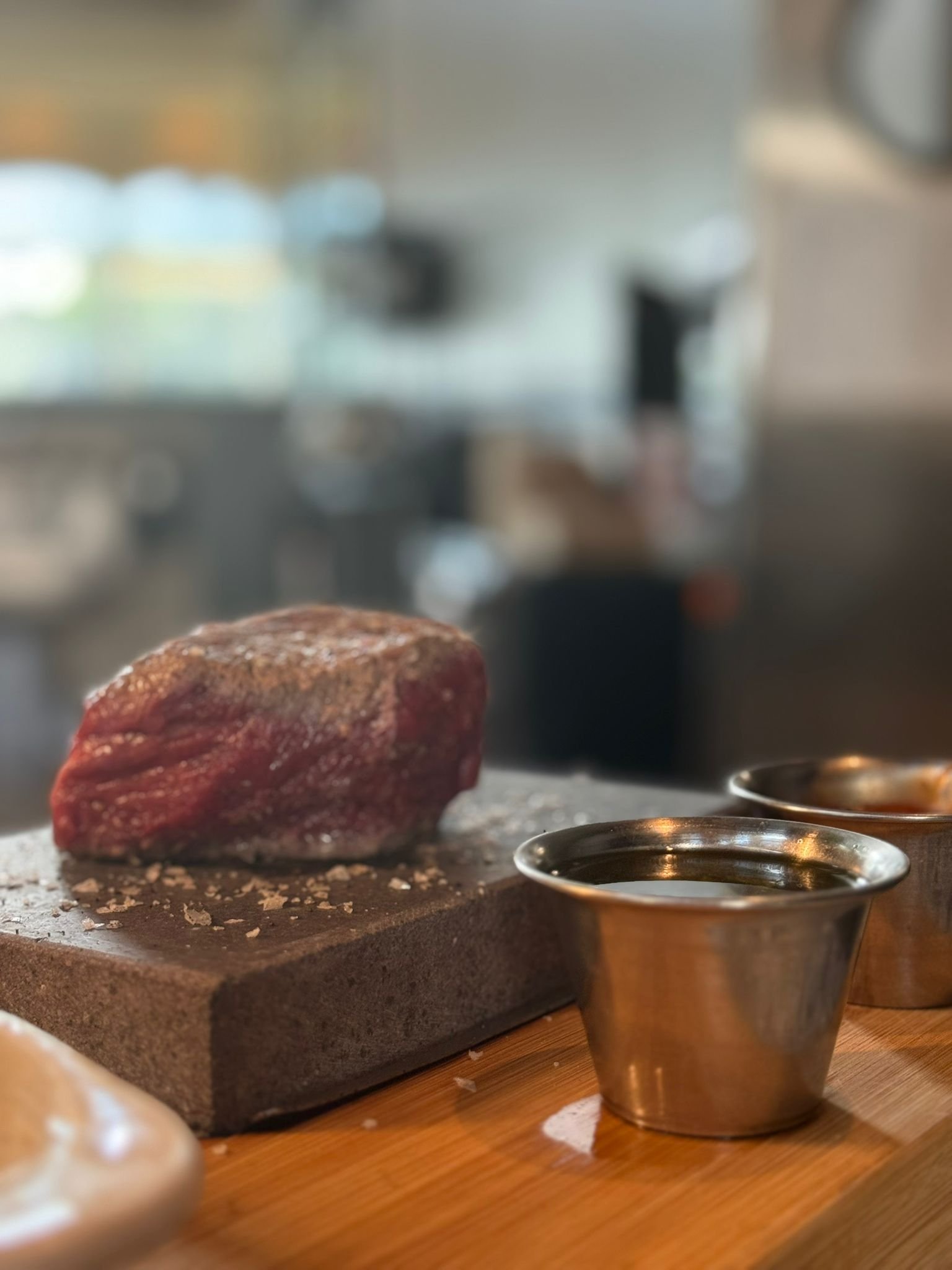 Raw piece of meat on a cutting board with salt, next to metal sauce cups on a wooden surface.