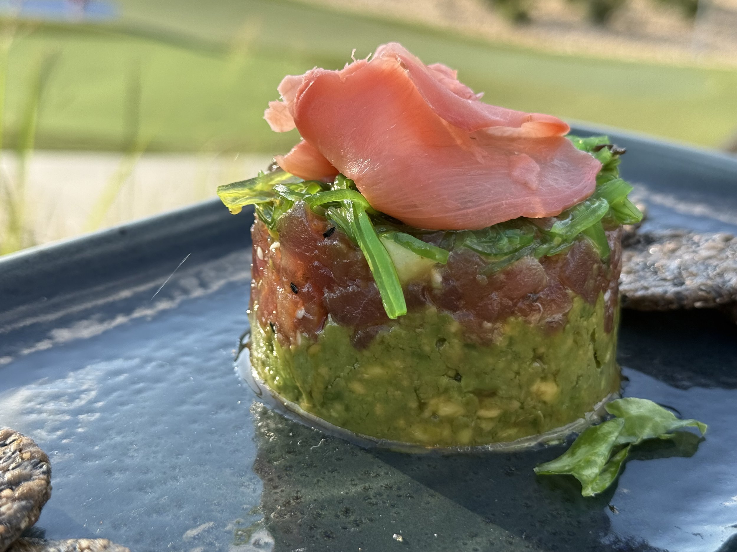 A close-up of a gourmet tuna tartare plated with avocado, topped with thin slices of cured ham and garnished with chopped herbs, served on a dark plate outdoors.