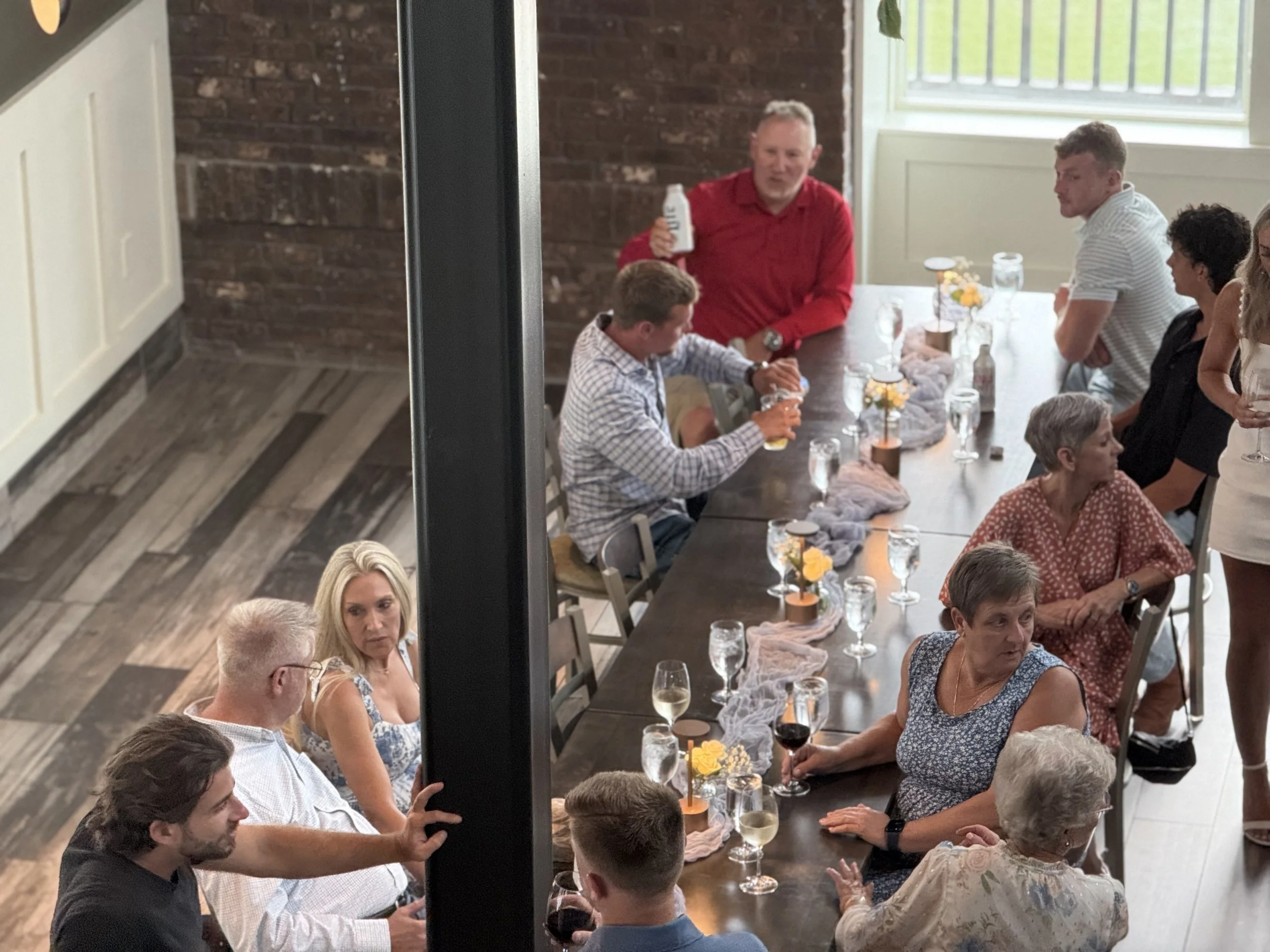 People gathered around a long dining table at a casual celebration or gathering, enjoying drinks and conversations in a well-lit room with brick walls and wooden flooring.