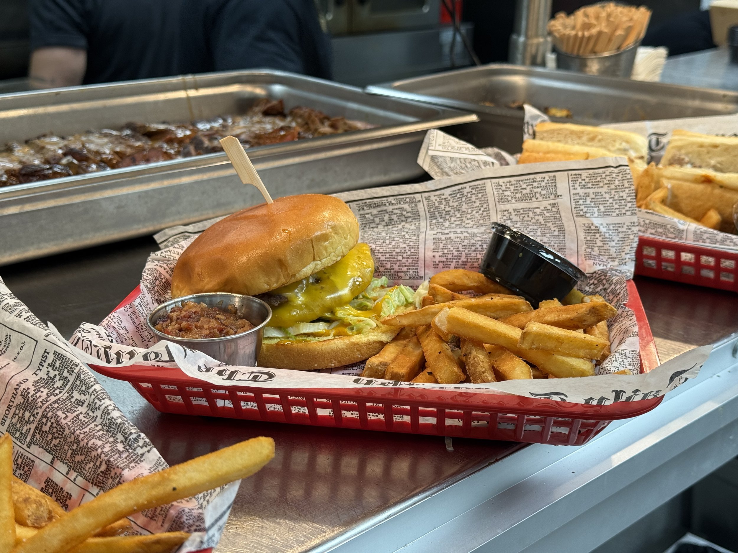 A cheeseburger with pickles on a bun, served with French fries and a small cup of chili in a red basket lined with newspaper print. In the background, there are trays of other food items, including sandwiches and skewers.