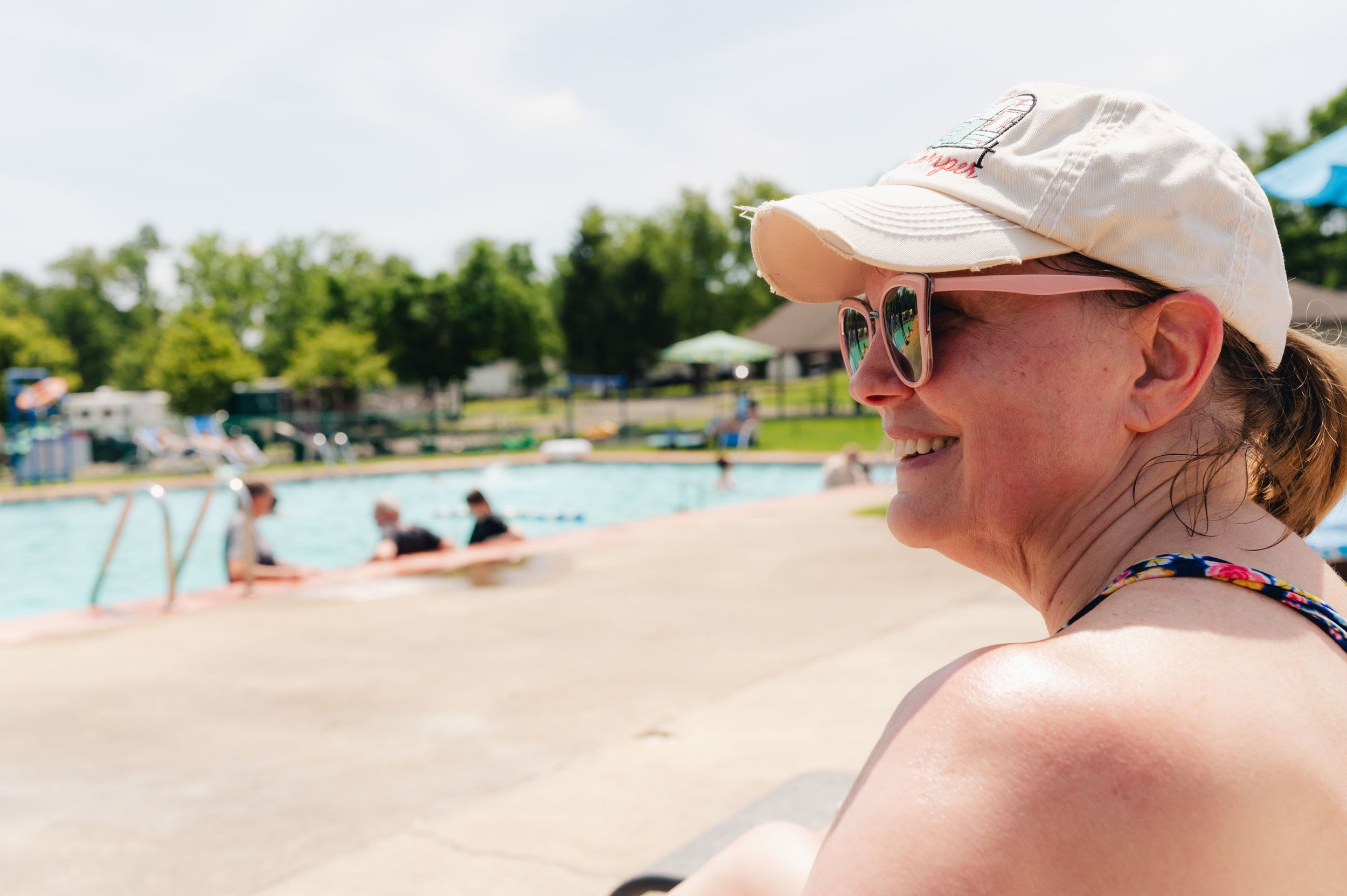 Smiling woman with sunglasses and a baseball cap sitting by a swimming pool on a sunny day, with people swimming and relaxing in the background.