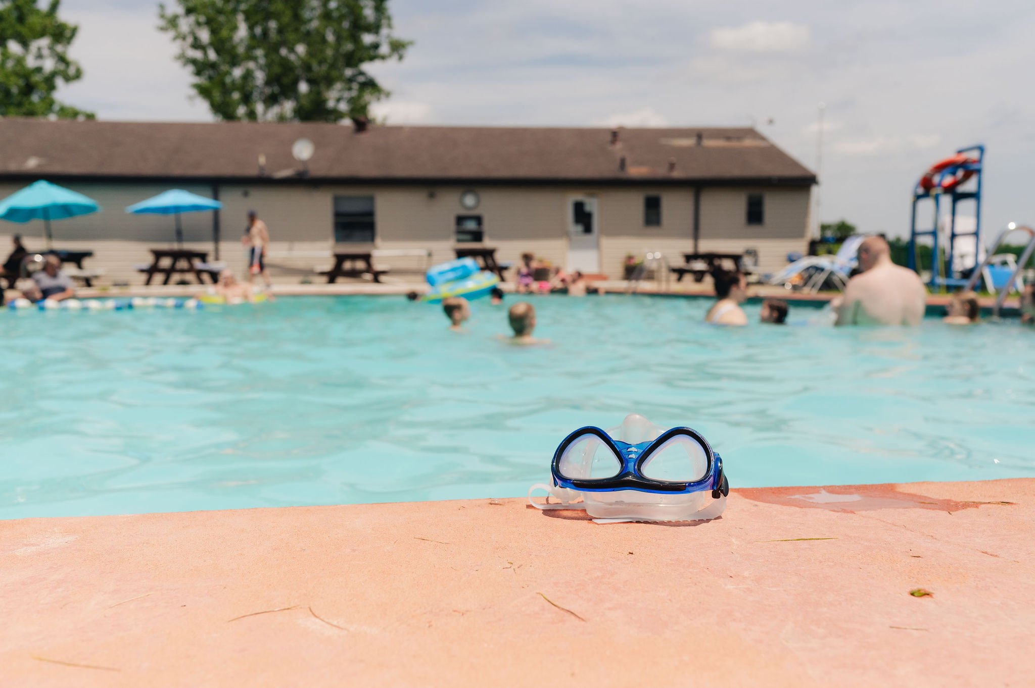 Swimming goggles on poolside with people swimming and relaxing in a backyard swimming pool, with patio tables and umbrellas in the background.