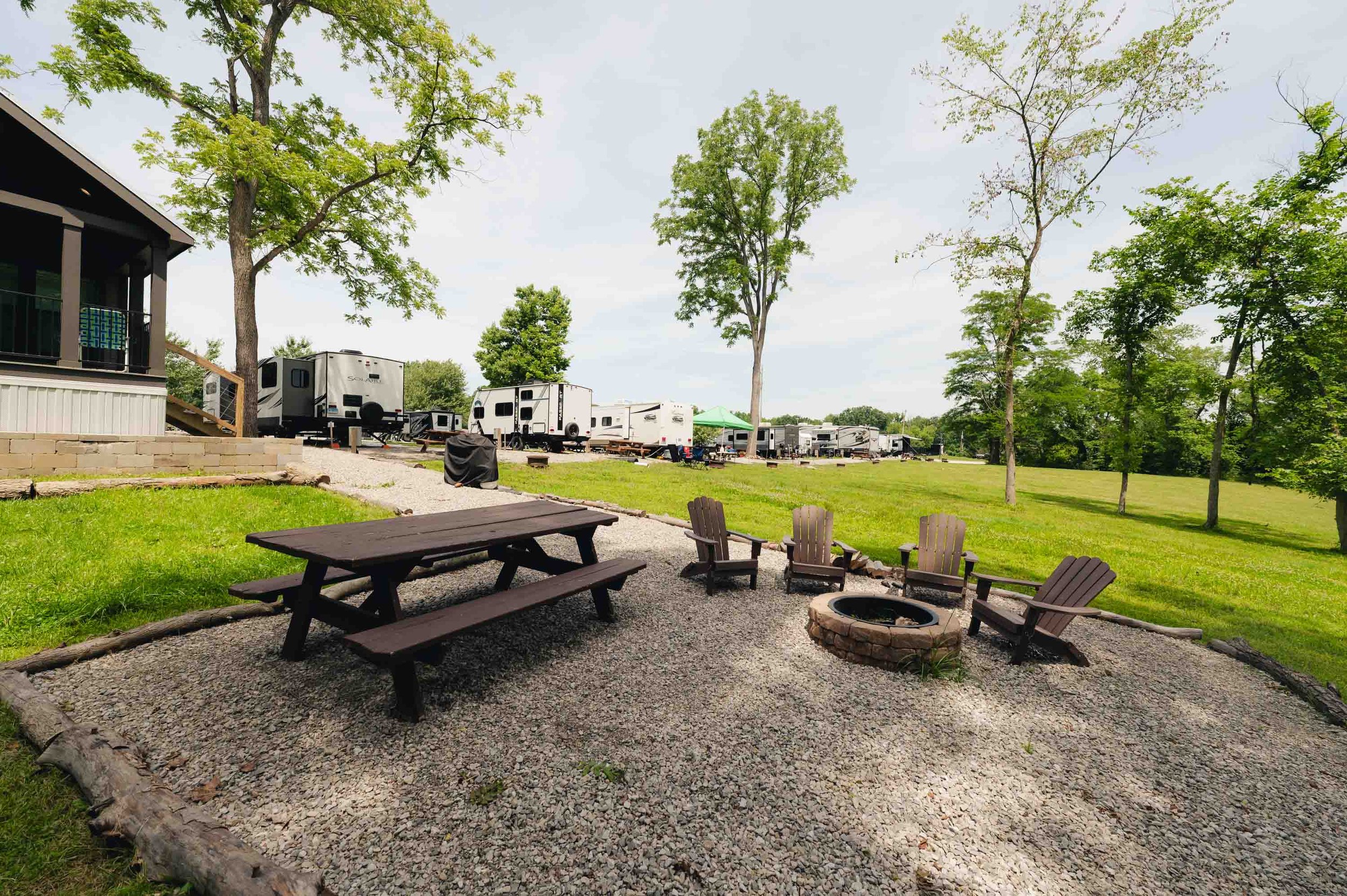 Outdoor scene with a gravel seating area featuring a picnic table and five Adirondack chairs surrounding a fire pit, with RVs parked in the background on a grassy lot, trees scattered around, and a building with a porch on the left.
