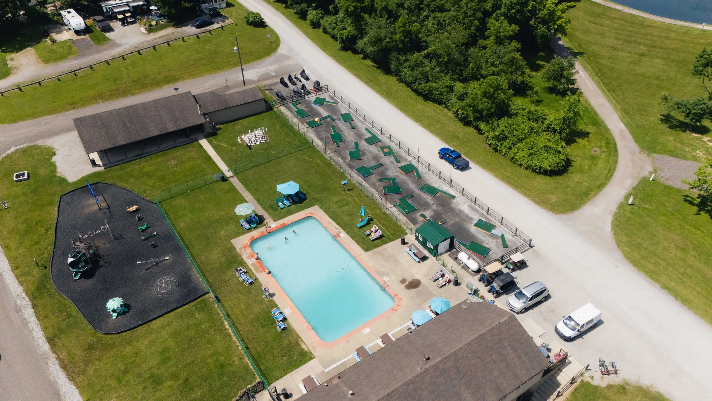 Aerial view of a recreational park area including a swimming pool with people, a playground, a corn hole game area, shaded tables, parking spaces with vehicles, and surrounding greenery and pathways.