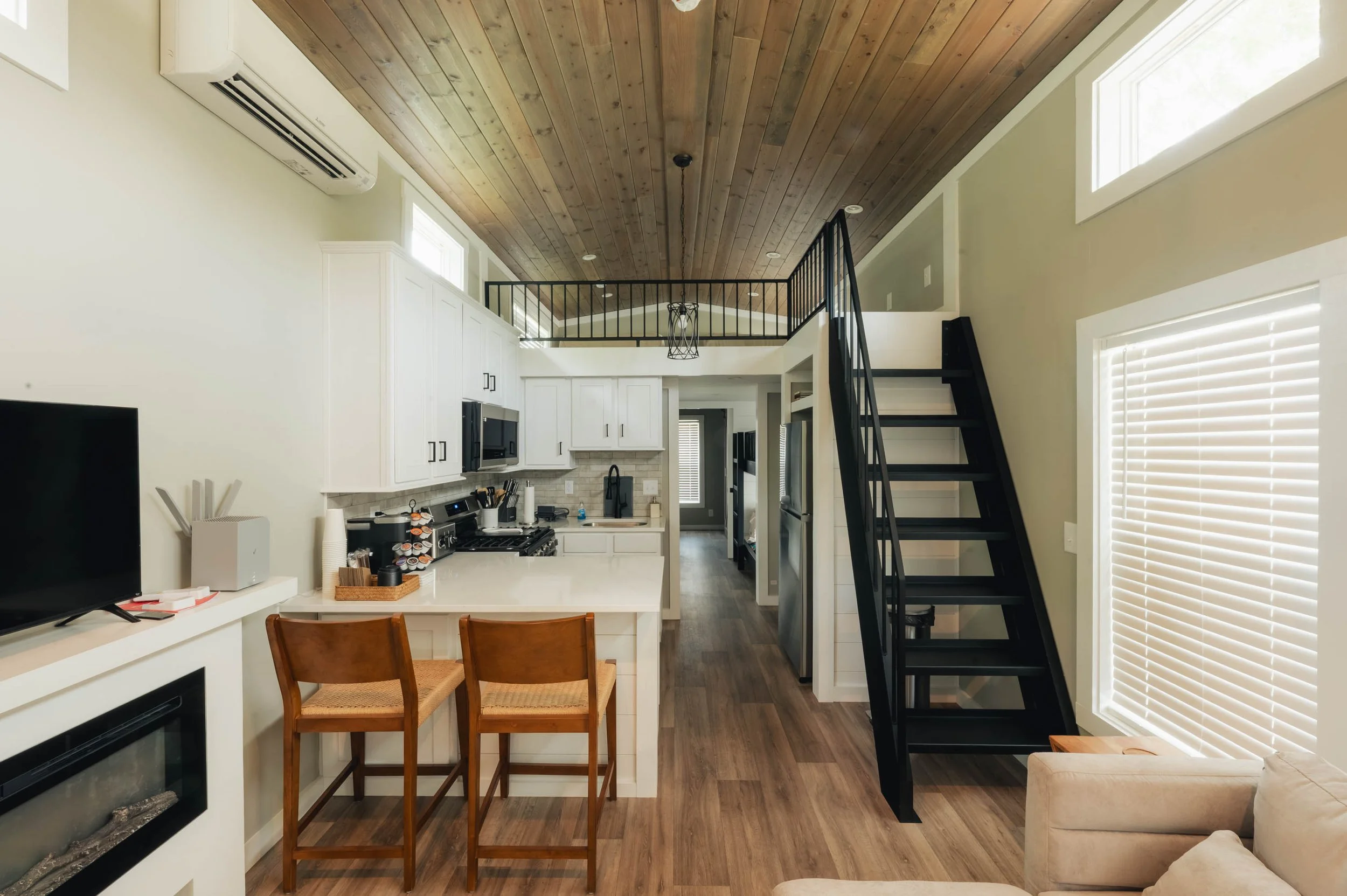 Open-concept living space with kitchen, dining area, and loft, featuring wood ceiling, black staircase, and large windows.
