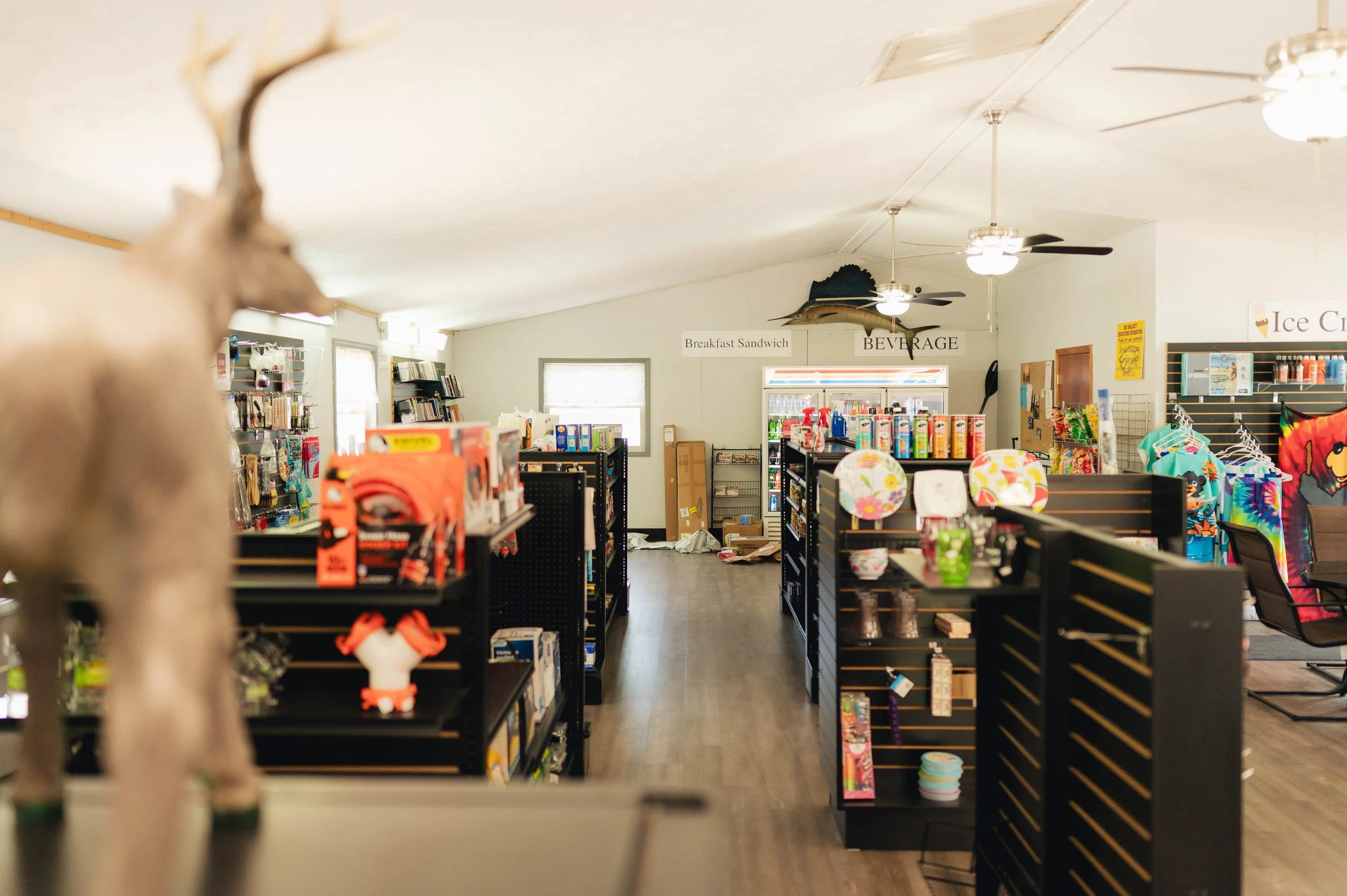 Interior of a store with shelves filled with various products, signs indicating 'Breakfast Sandwich' and 'Beverage', ceiling fans, and a corner with clothing and souvenirs. The store has wooden floors and natural light from windows.