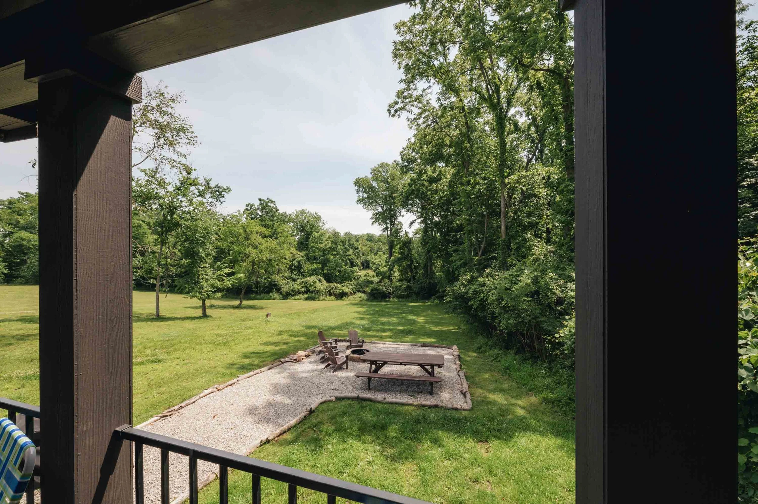 View of a backyard seen from a porch, showing green grass, a gravel sitting area with wooden chairs and table, surrounded by trees and shrubs under a partly cloudy sky.