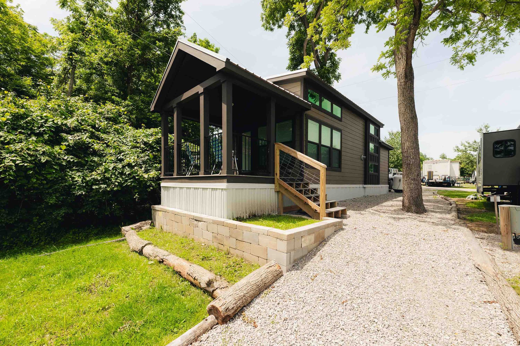 A modern tiny house on a raised foundation with a porch and a set of stairs, surrounded by green trees and a gravel pathway.