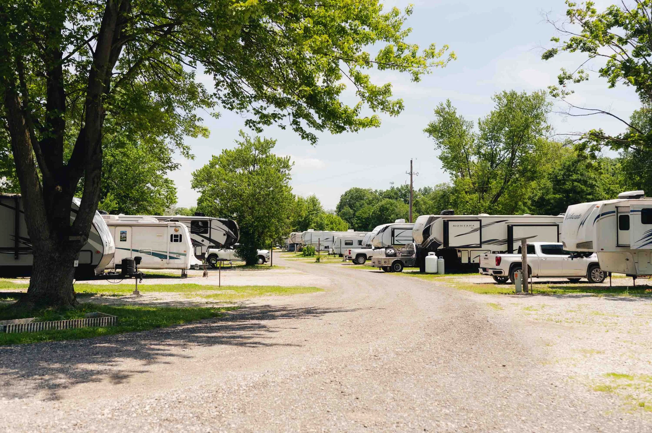 A campground with multiple recreational vehicles parked along a gravel road under green trees on a sunny day.