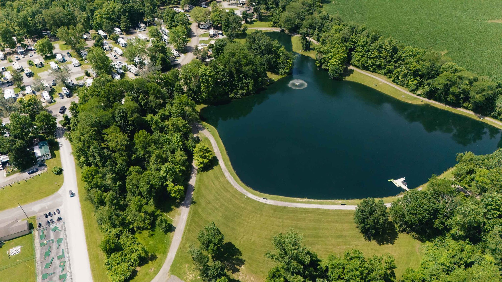 Aerial view of a park with a pond, fountain, sandy beach, trees, grassy areas, picnic tables, and a parking lot with many RVs and vehicles.