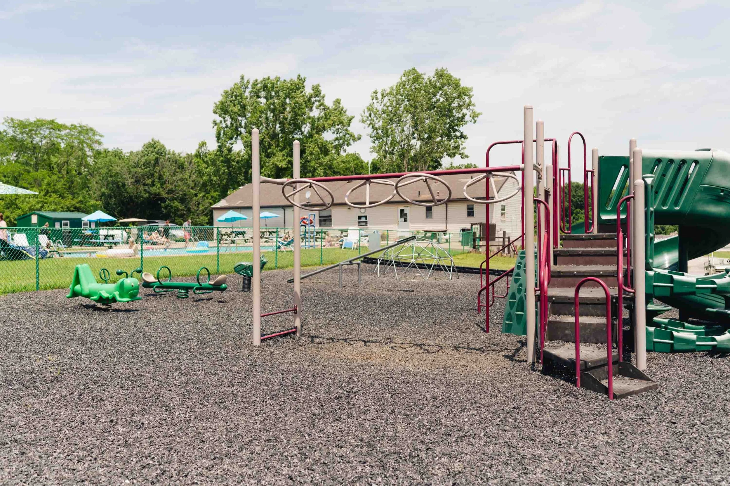 Empty playground with climbing structures, slides, and spring riders, next to a swimming pool area with umbrellas and outdoor seating.