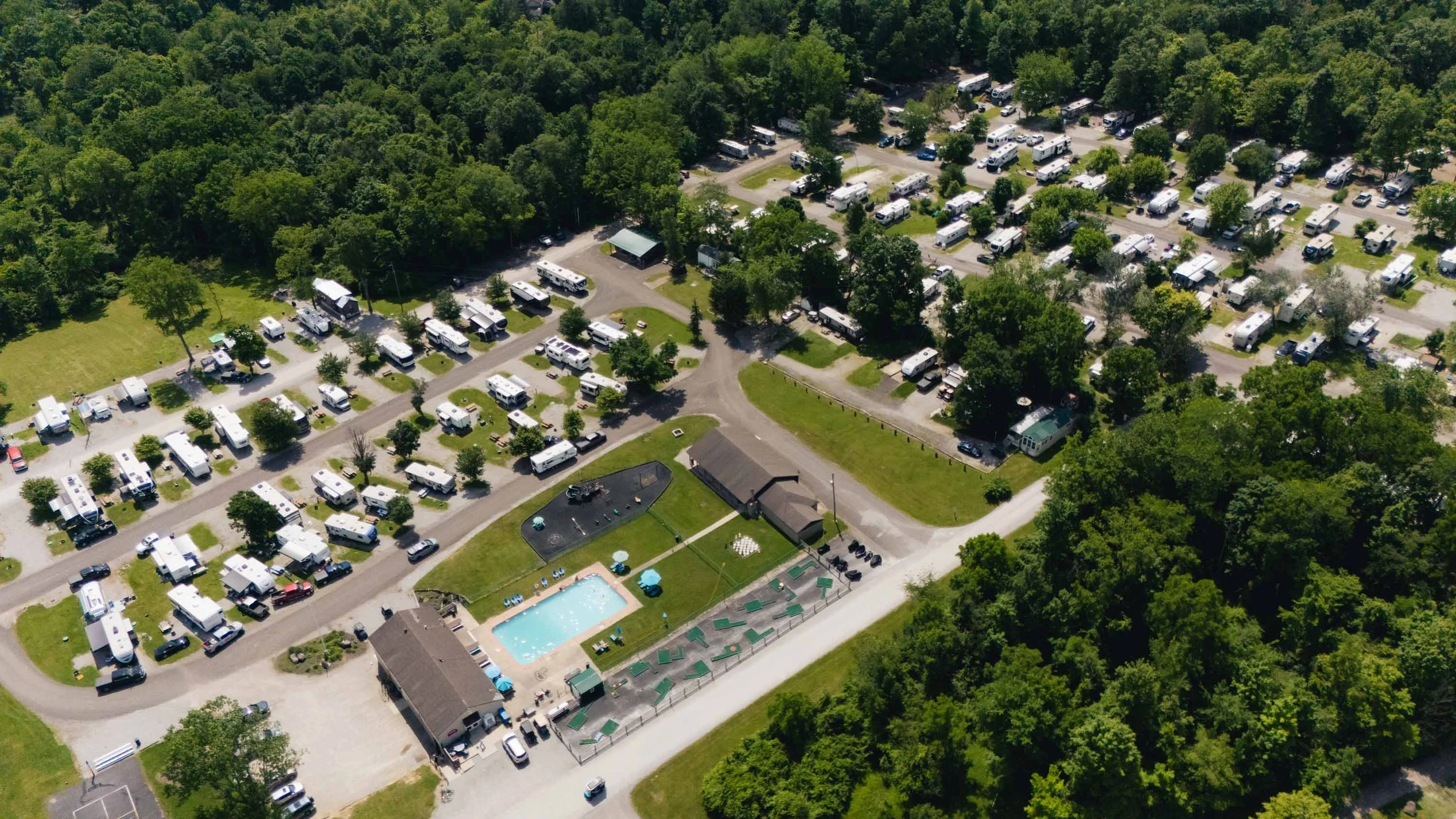 Aerial view of a campground with numerous RVs and trailers parked among trees, a swimming pool, playground, and a small building, surrounded by lush green forest.