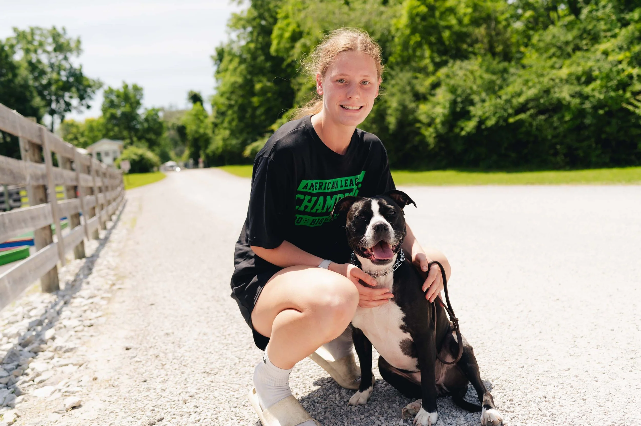 A young woman kneeling on a gravel path outdoors, smiling and holding a black and white dog with a chain collar, with greenery in the background.