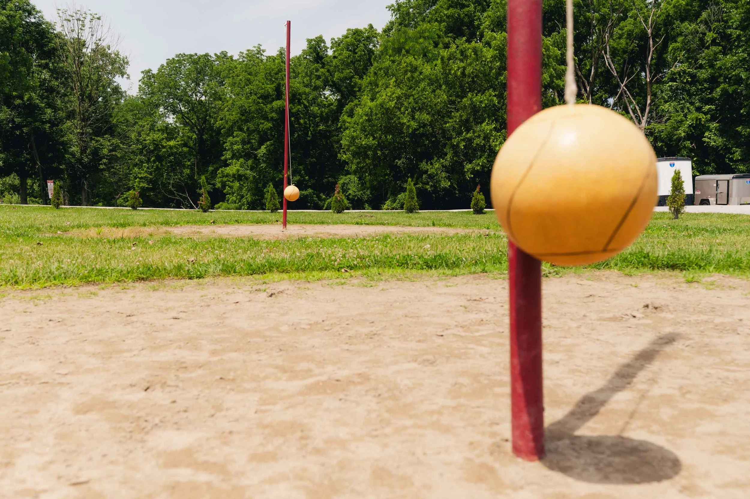 Close-up of two wooden balls hanging on red poles in a park with grass, trees, and parked buses in the background.