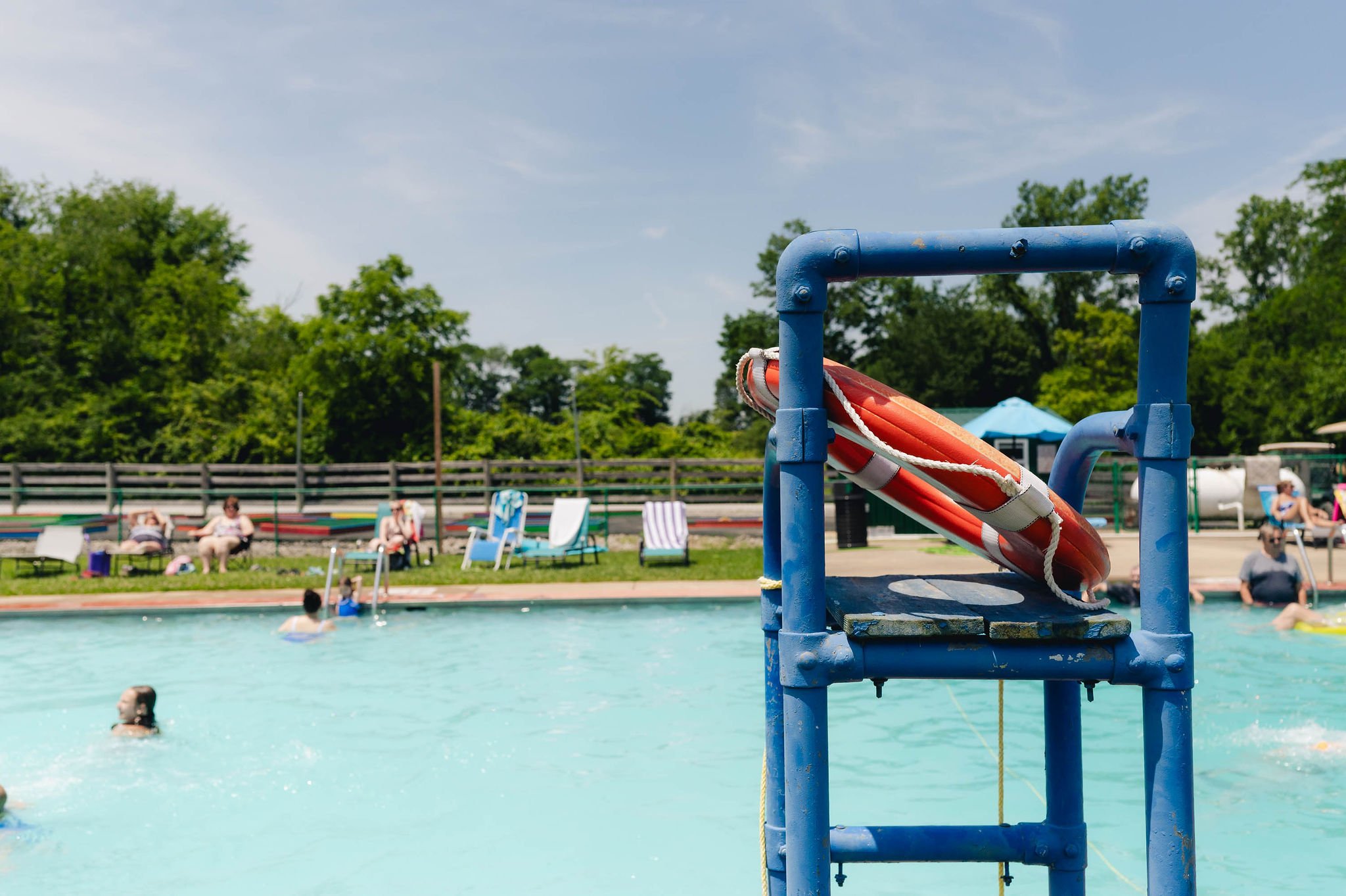 A lifebuoy on a blue lifeguard stand at an outdoor swimming pool, with people swimming and lounging on chairs in the background, and green trees under a partly cloudy sky.