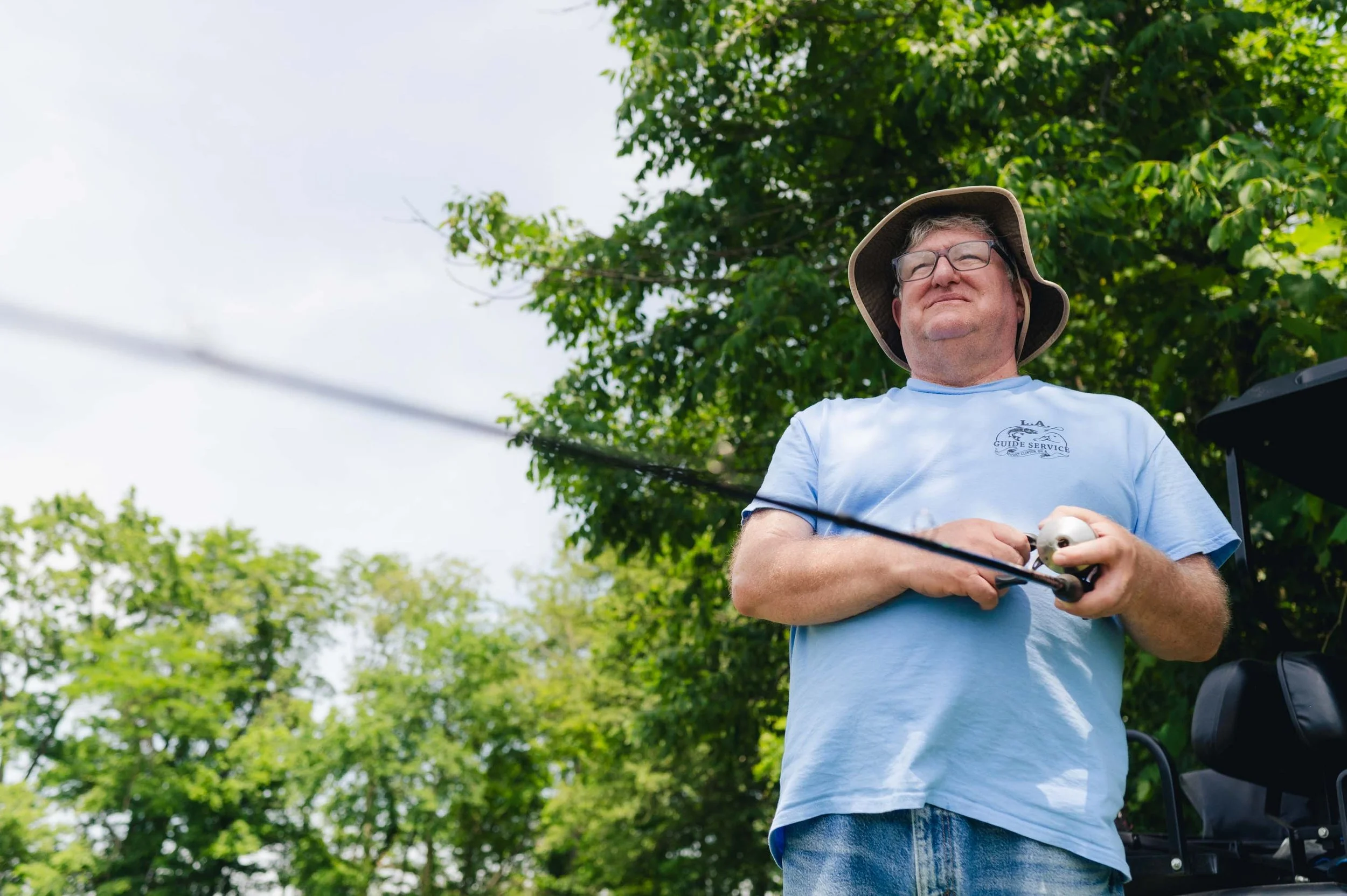 A man wearing glasses, a sun hat, and a light blue T-shirt standing outdoors with green trees in the background, holding a remote control for a drone.