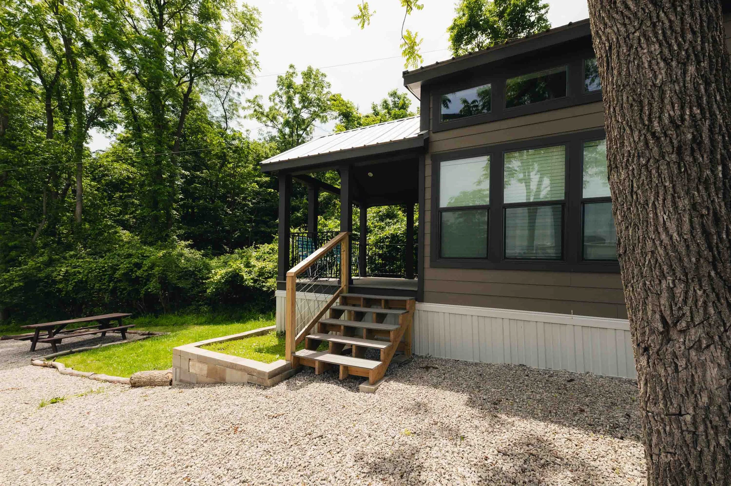 Exterior of a modern house with a small wooden porch, steps, and black window frames, situated in a lush, green area with trees and a gravel pathway.