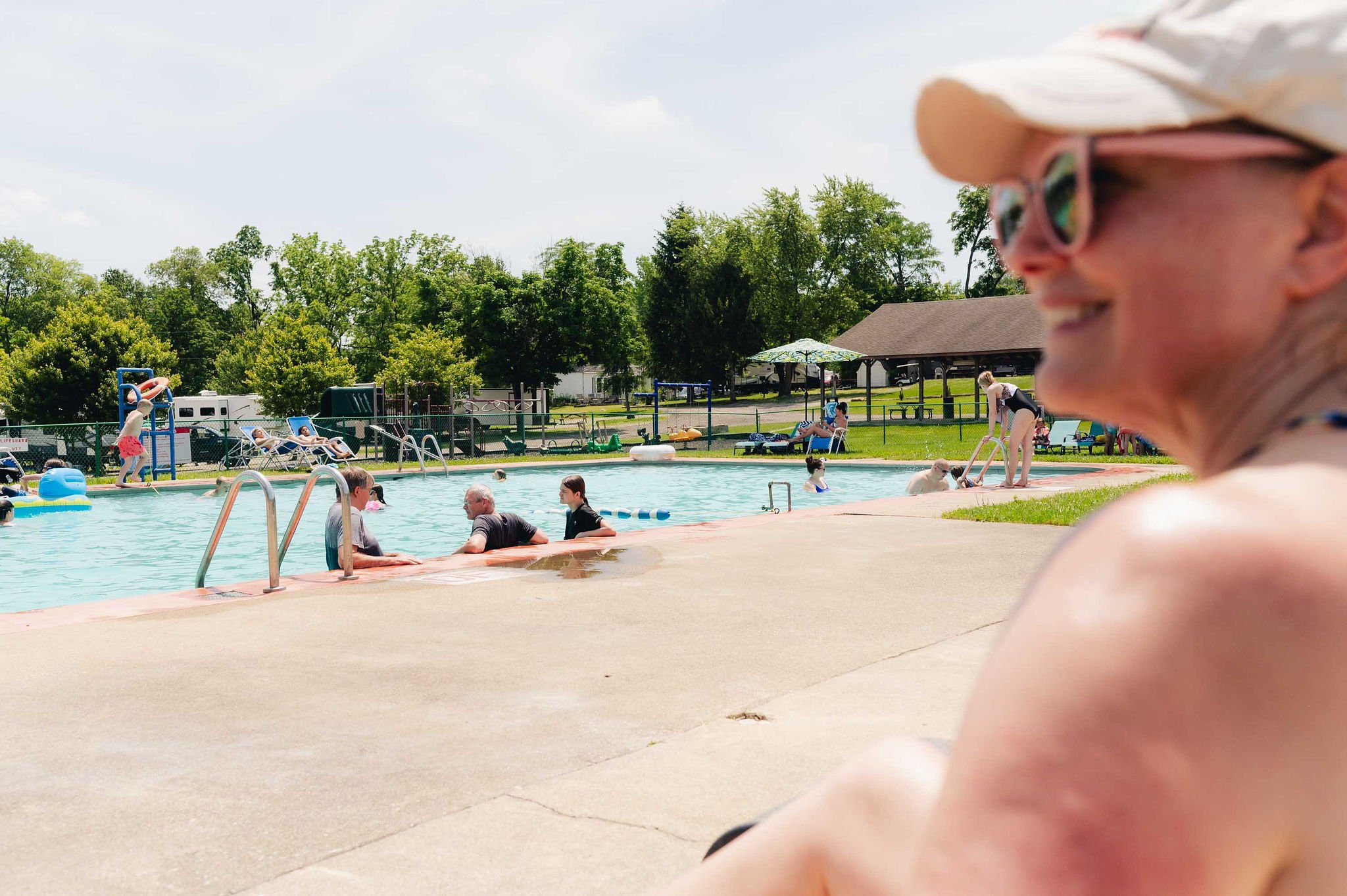 A woman with glasses and a hat sitting near a swimming pool on a sunny day, with children playing and adults relaxing in and around the pool.