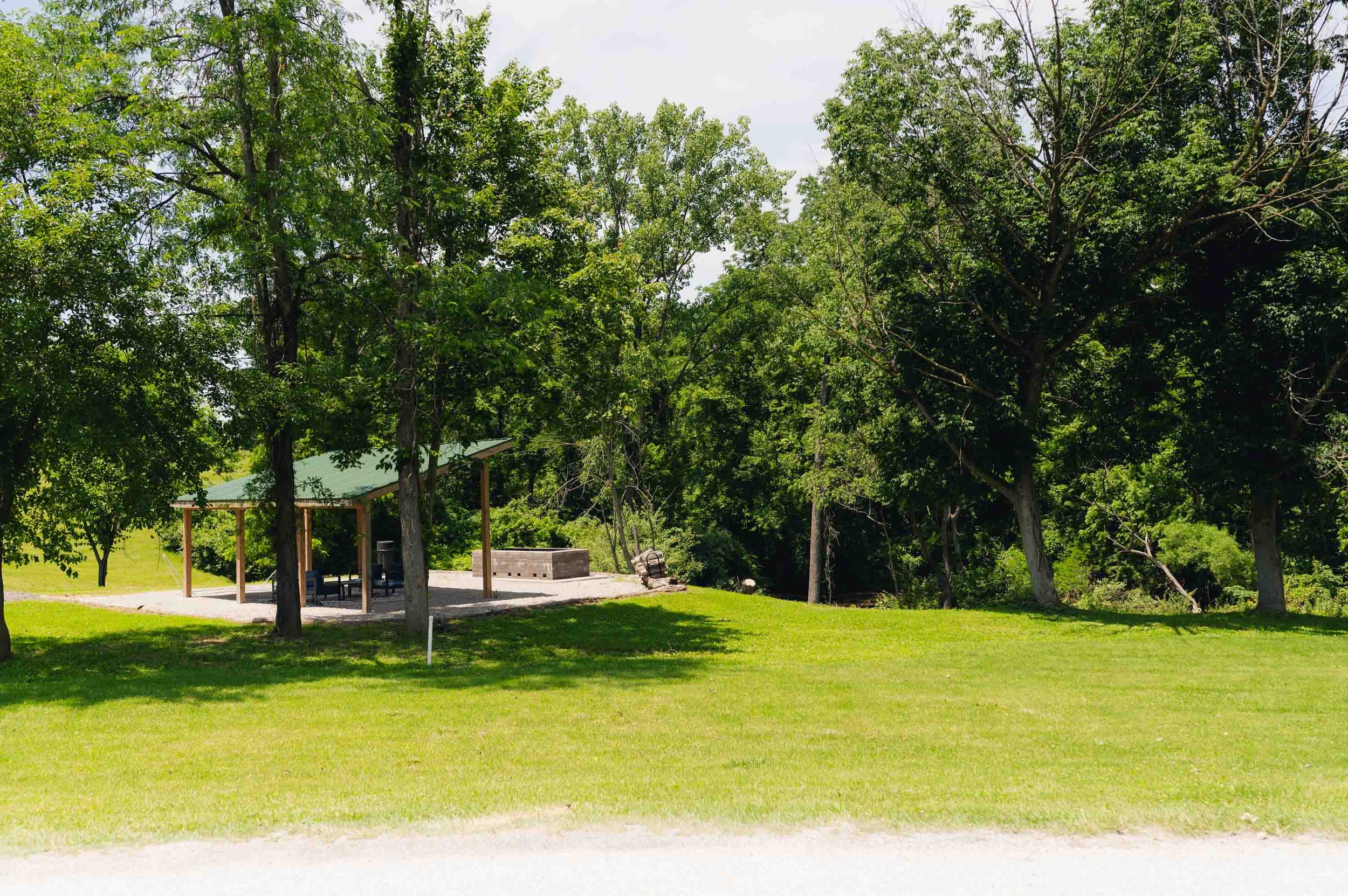 A park with a grassy lawn, several large trees, a covered picnic area with tables, and a stone fire pit, under a partly cloudy sky.