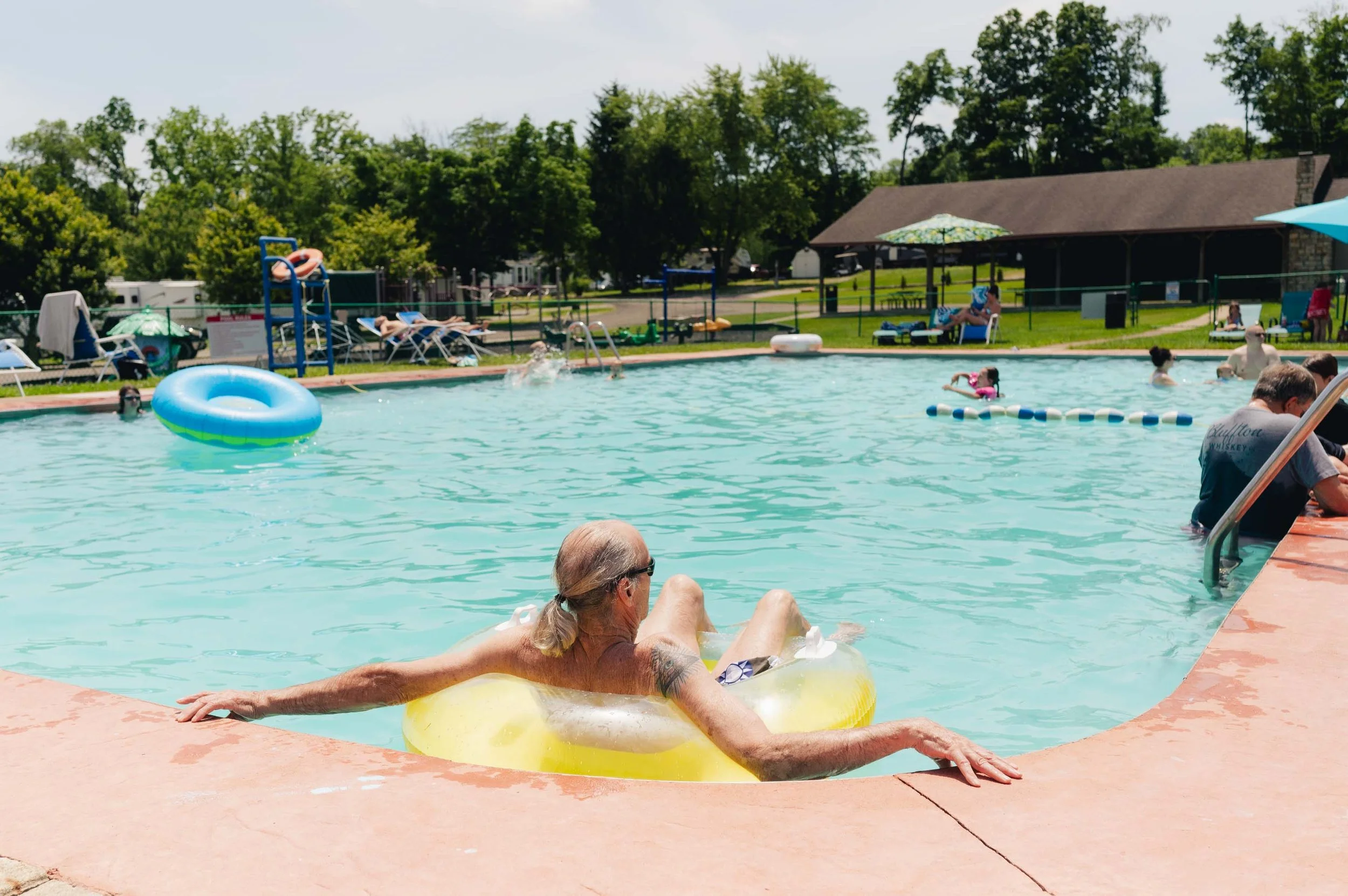 A swimming pool with people enjoying a sunny day, including an elderly woman with gray hair in sunglasses relaxing on a yellow float near the edge of the pool, children swimming and playing, and lounge chairs with umbrellas surrounded by green trees 