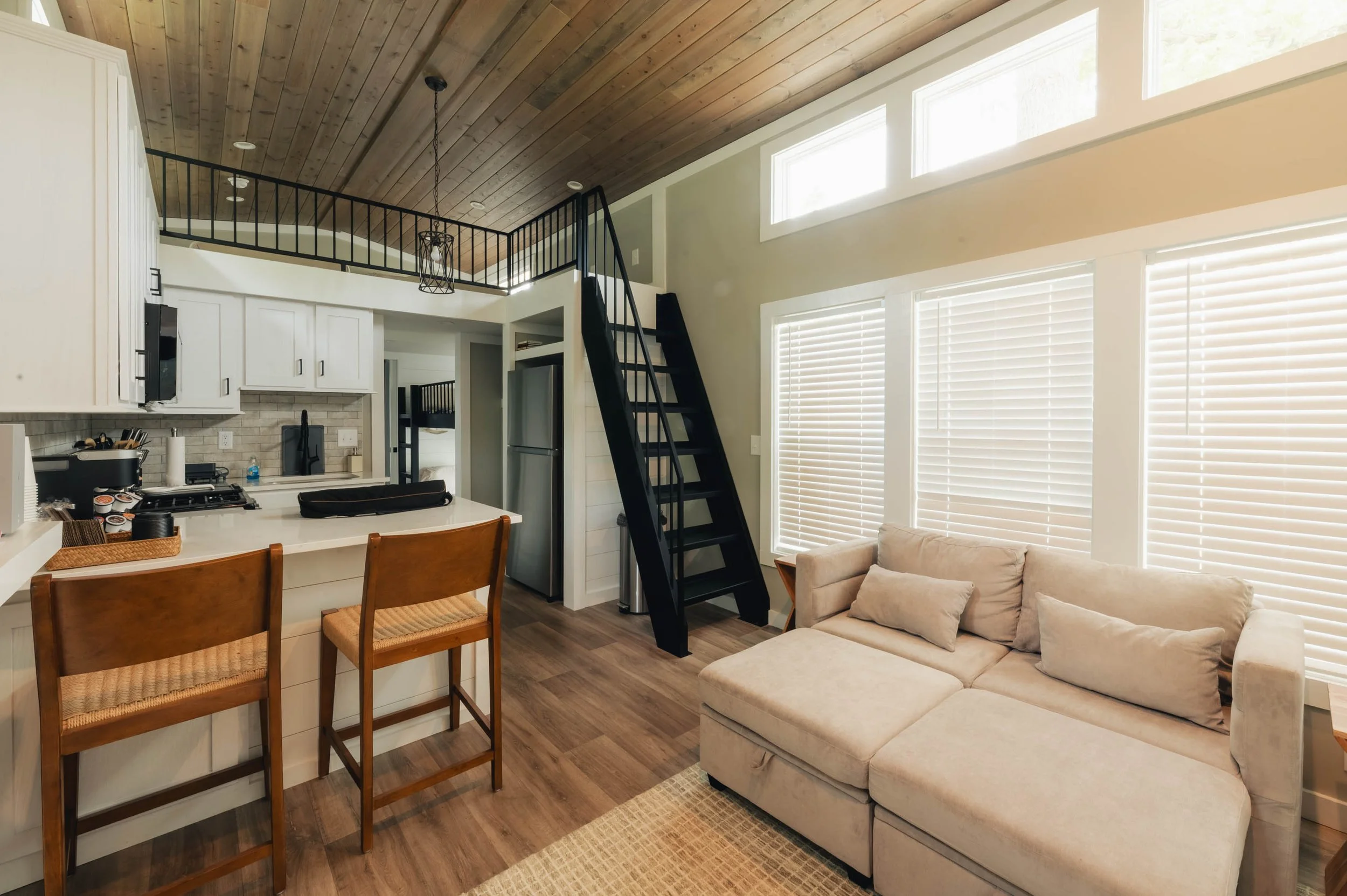 Living room and kitchen area with wood ceiling, black staircase, white cabinets, beige couch, and large windows with blinds.