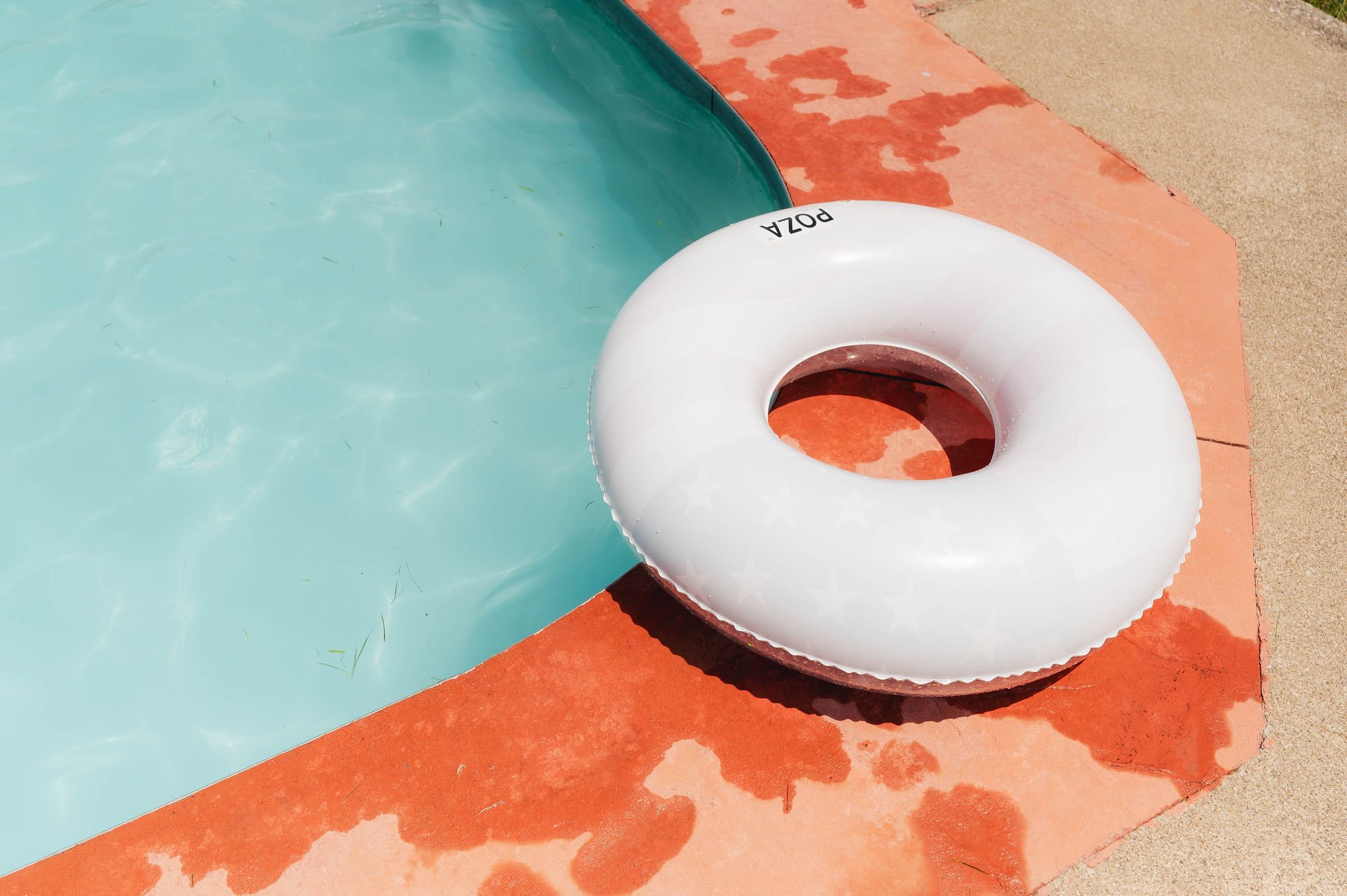 An inflatable white swim ring labeled 'POZA' resting on the edge of a swimming pool with light blue water, on a wet reddish-orange concrete surface.